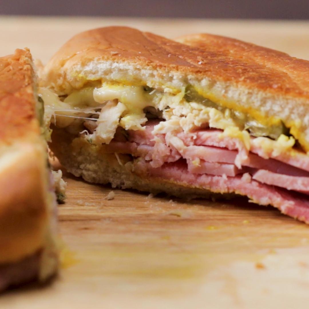 Cuban sandwich on a wooden board, showcasing layers of ham, roast pork, Swiss cheese, pickles, and mustard between crispy bread