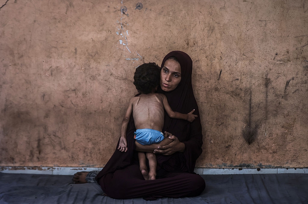 A woman in a seated position holds a child who is standing. Both have a somber expression. The wall behind them is textured and worn