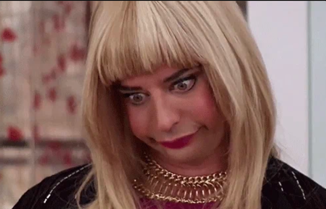 Person med overdrevet uttrykk, iført en blond parykk, rød leppestift og et gullkjede, og etterligner en komisk stil