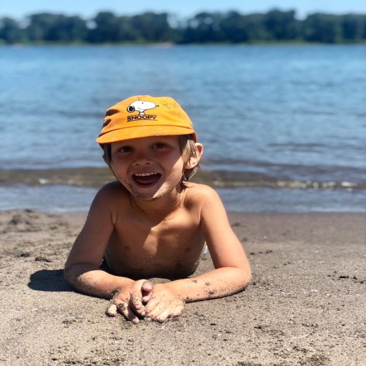 Child smiling while lying on sandy beach near water, wearing a cap with cartoon character design