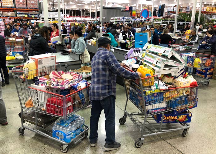 A crowded supermarket with a person pushing carts filled with groceries, including water and snacks, amidst busy checkout lines