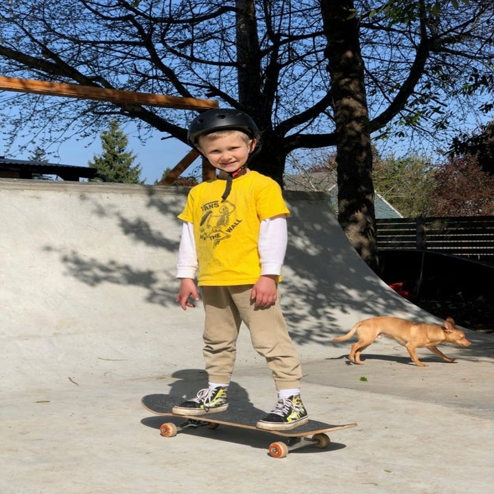 Child skateboarding in park, wearing helmet, yellow shirt, and beige pants. Small dog walking in the background