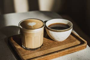 A latte with heart foam and a black coffee in a rustic bowl sit on a wooden tray