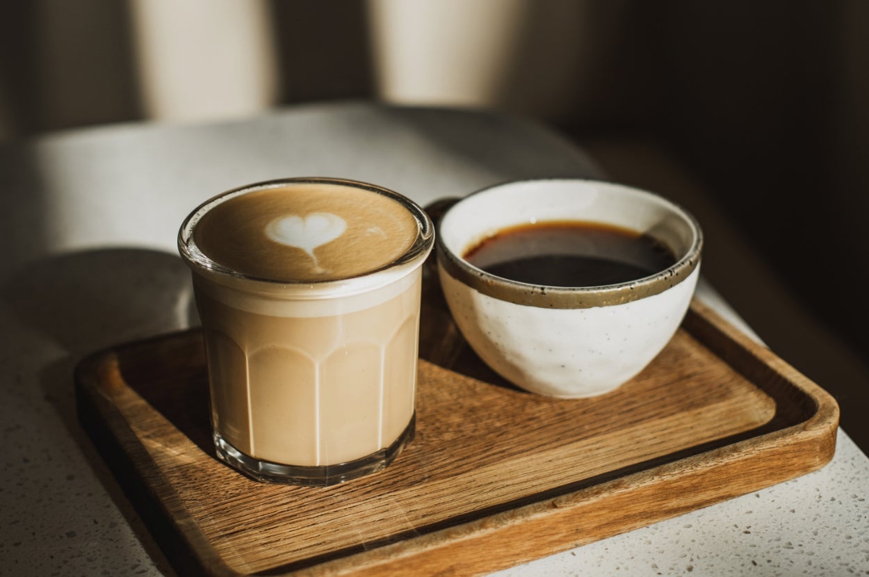 A latte with heart foam and a black coffee in a rustic bowl sit on a wooden tray