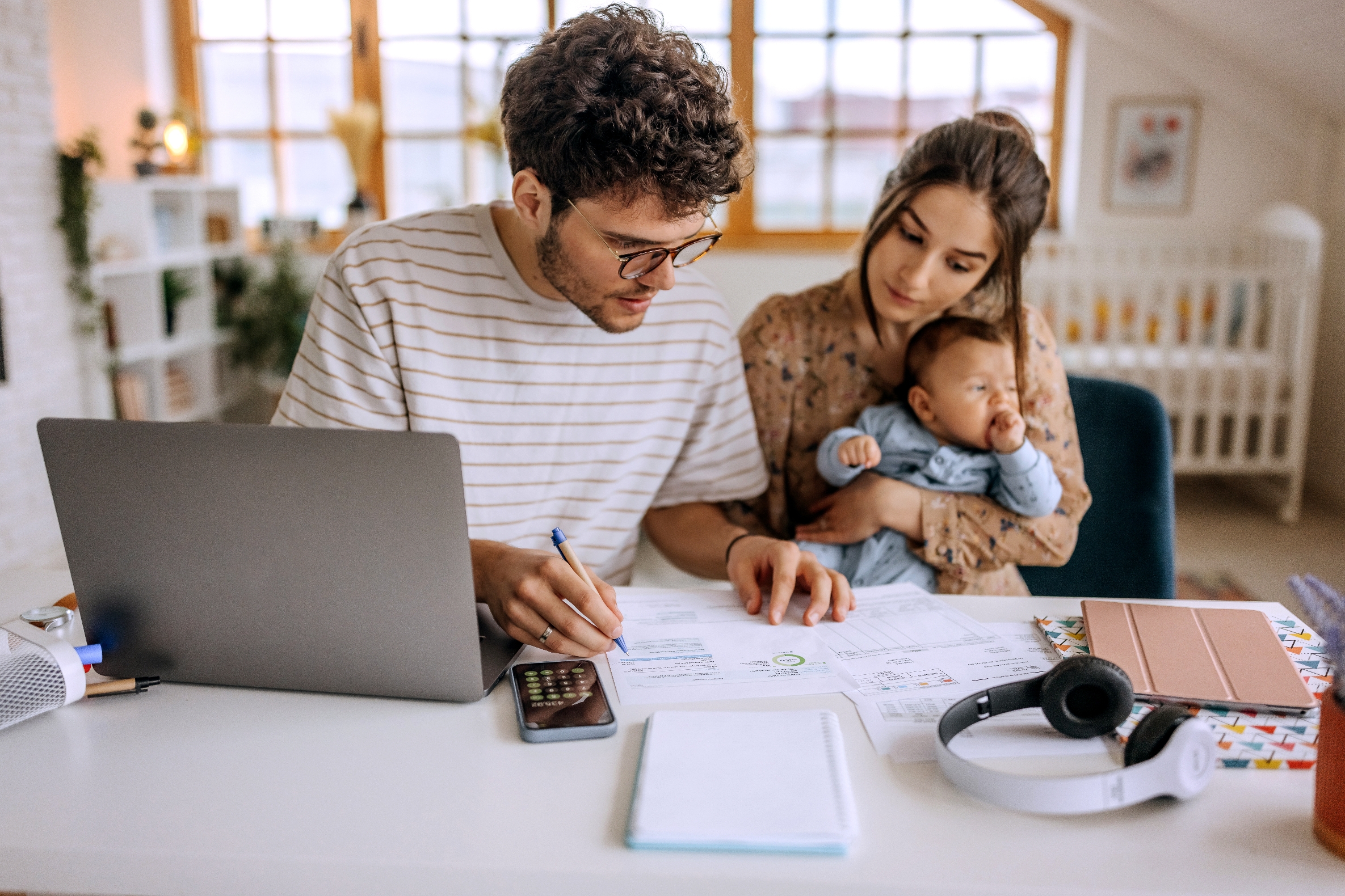 A couple sits at a desk with a baby, reviewing finances with a laptop, calculator, and documents, illustrating family financial planning