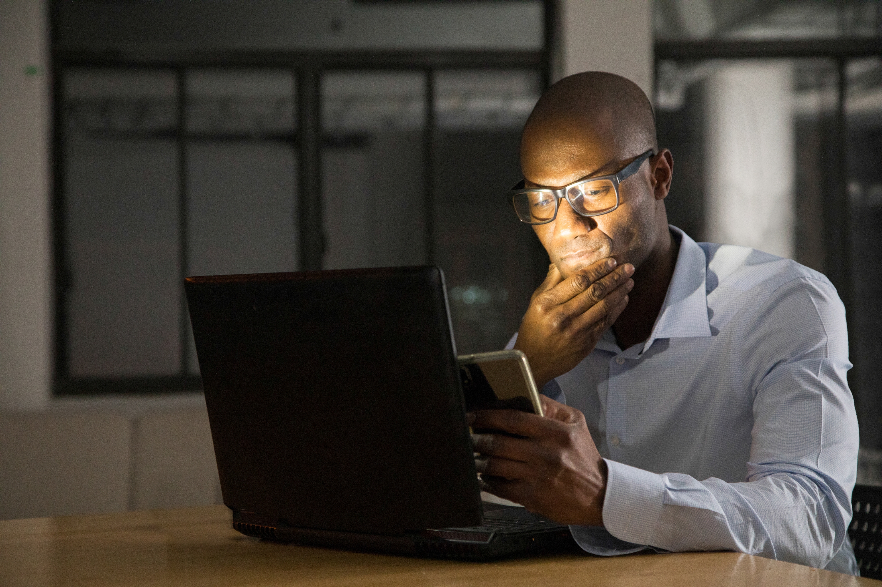 Person in formal attire looks at phone screen, sitting at a desk with a laptop. Office setting, suggesting work or business focus