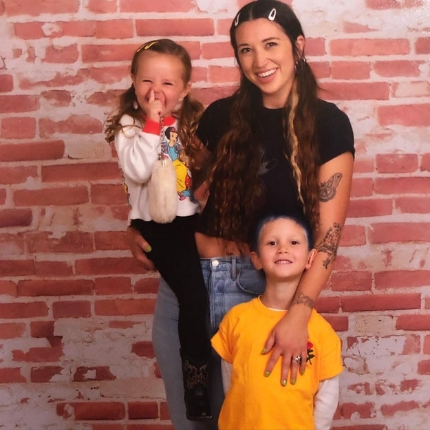 A smiling woman with long hair holds a laughing child while another child stands beside her against a brick wall backdrop