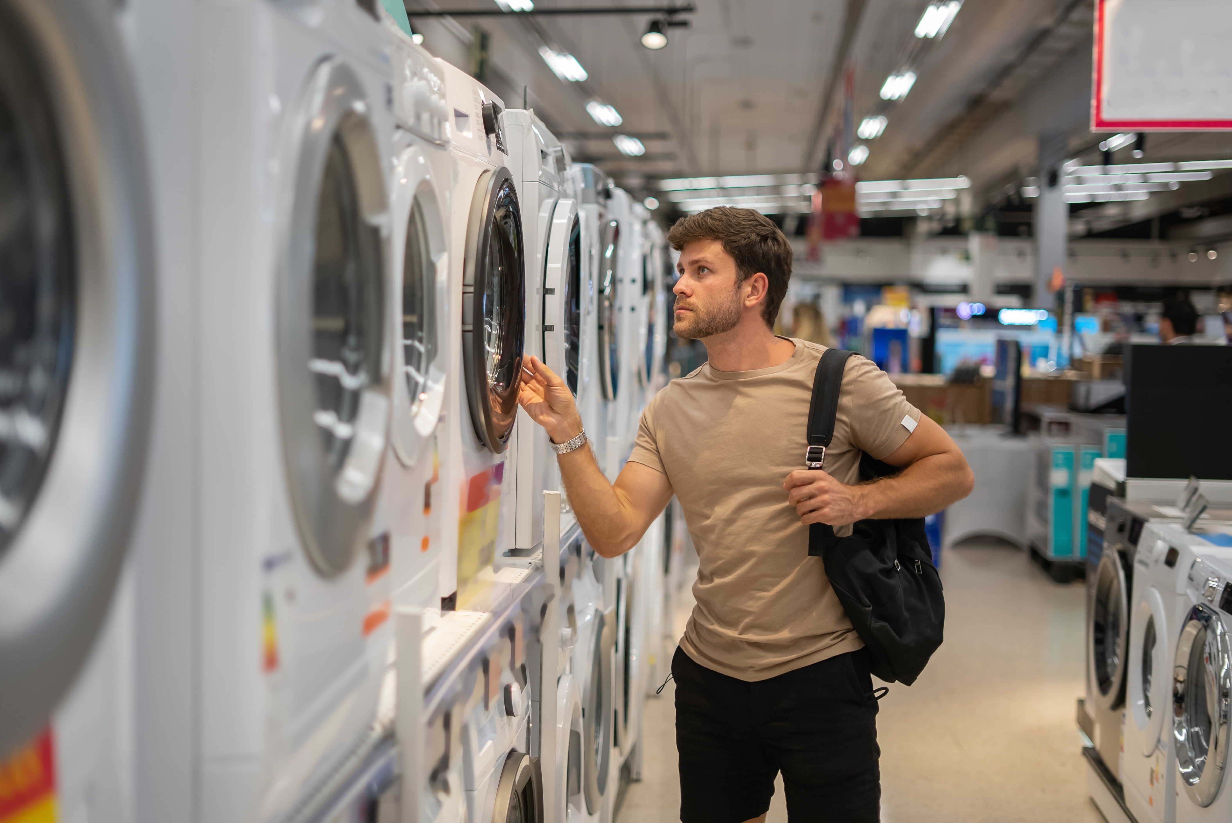 Man shopping for washing machines in an appliance store, examining displays thoughtfully with a backpack on his shoulder