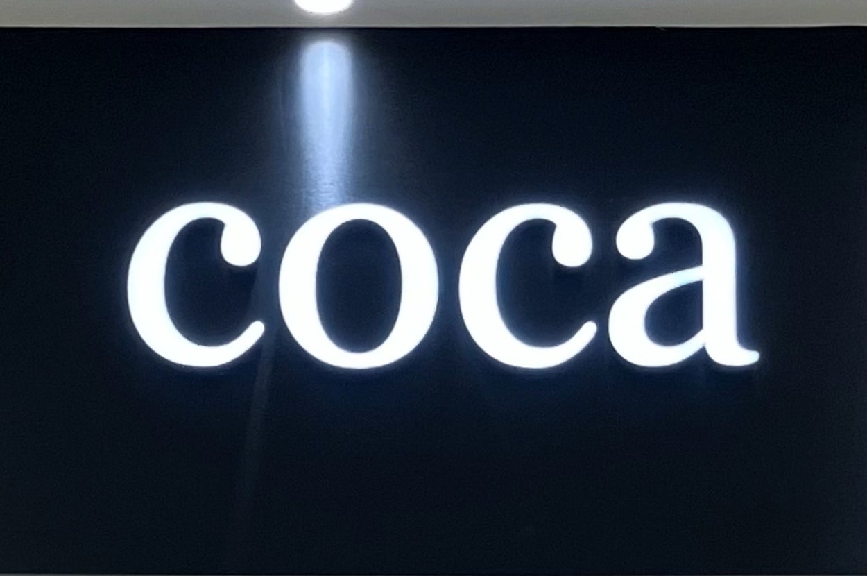 暗い背景に白い文字で「coca」と書かれた看板。店舗の入り口にある。