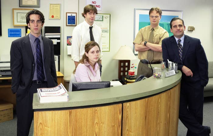 Five office workers, in business attire, gather around a reception desk. The setting is informal and reminiscent of a typical office environment