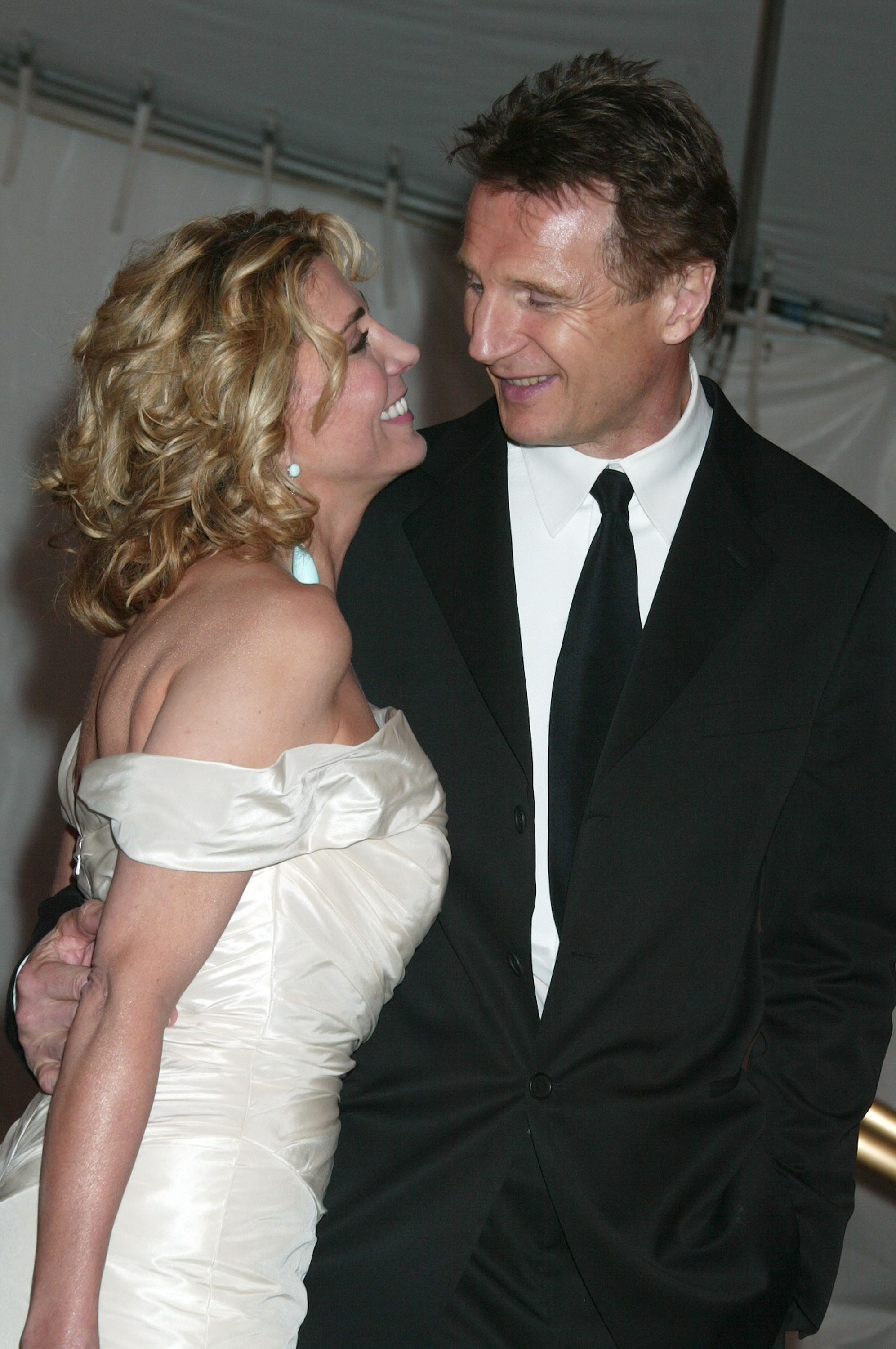 Two people smiling at each other on the red carpet. The woman is wearing an off-shoulder dress, and the man is in a suit and tie