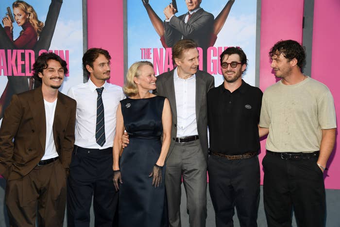 Group of six people smiling at a film premiere, with one person in a classic suit and others in casual wear. Movie posters visible in the background