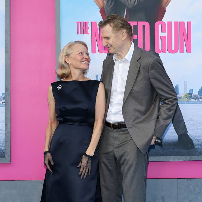 Two people smiling on a red carpet; one in a sophisticated black dress with gloves, the other in a tailored suit. A movie poster is in the background