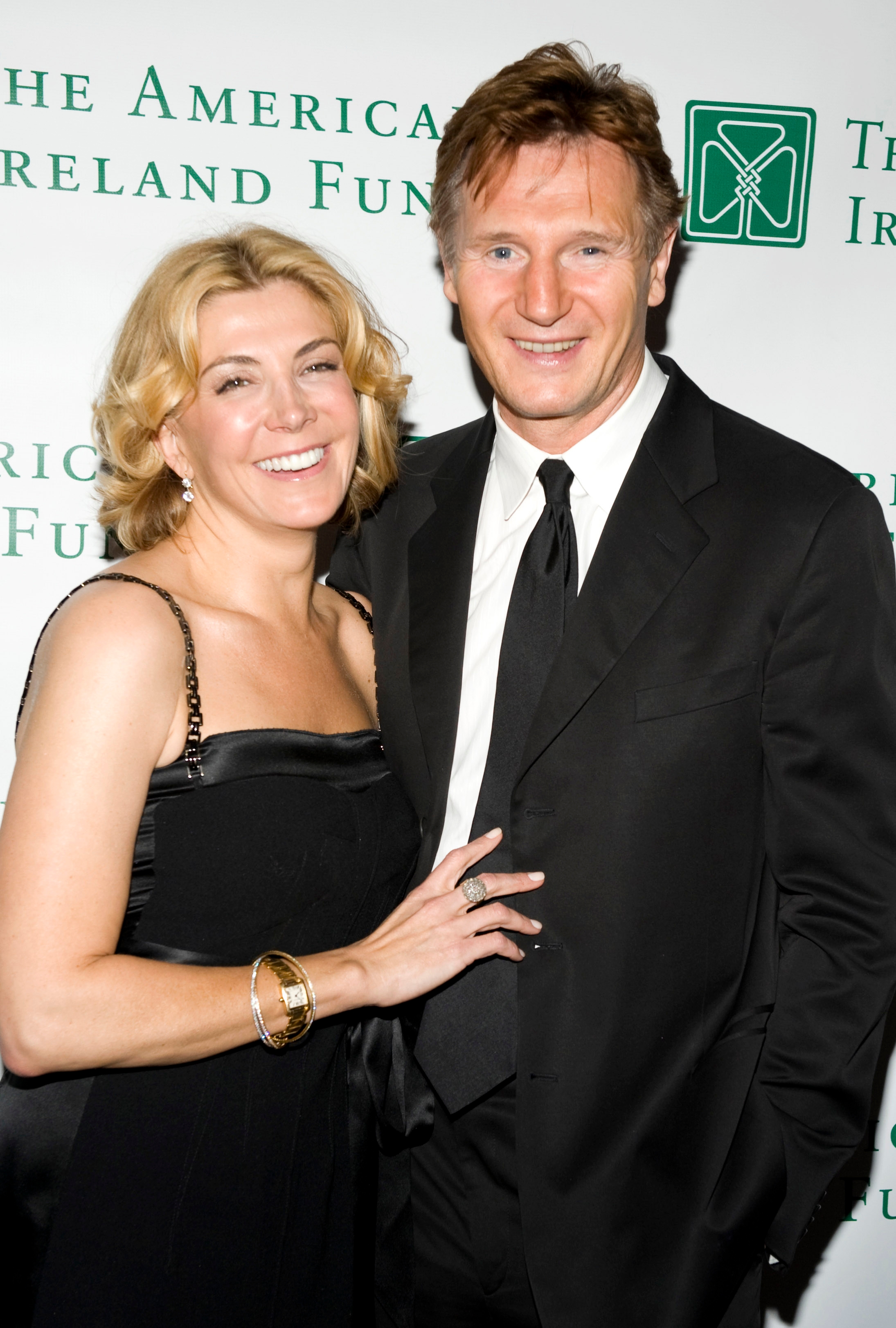 A woman in a stylish black strapless dress poses with a man in a classic black suit and tie at a formal event backdrop