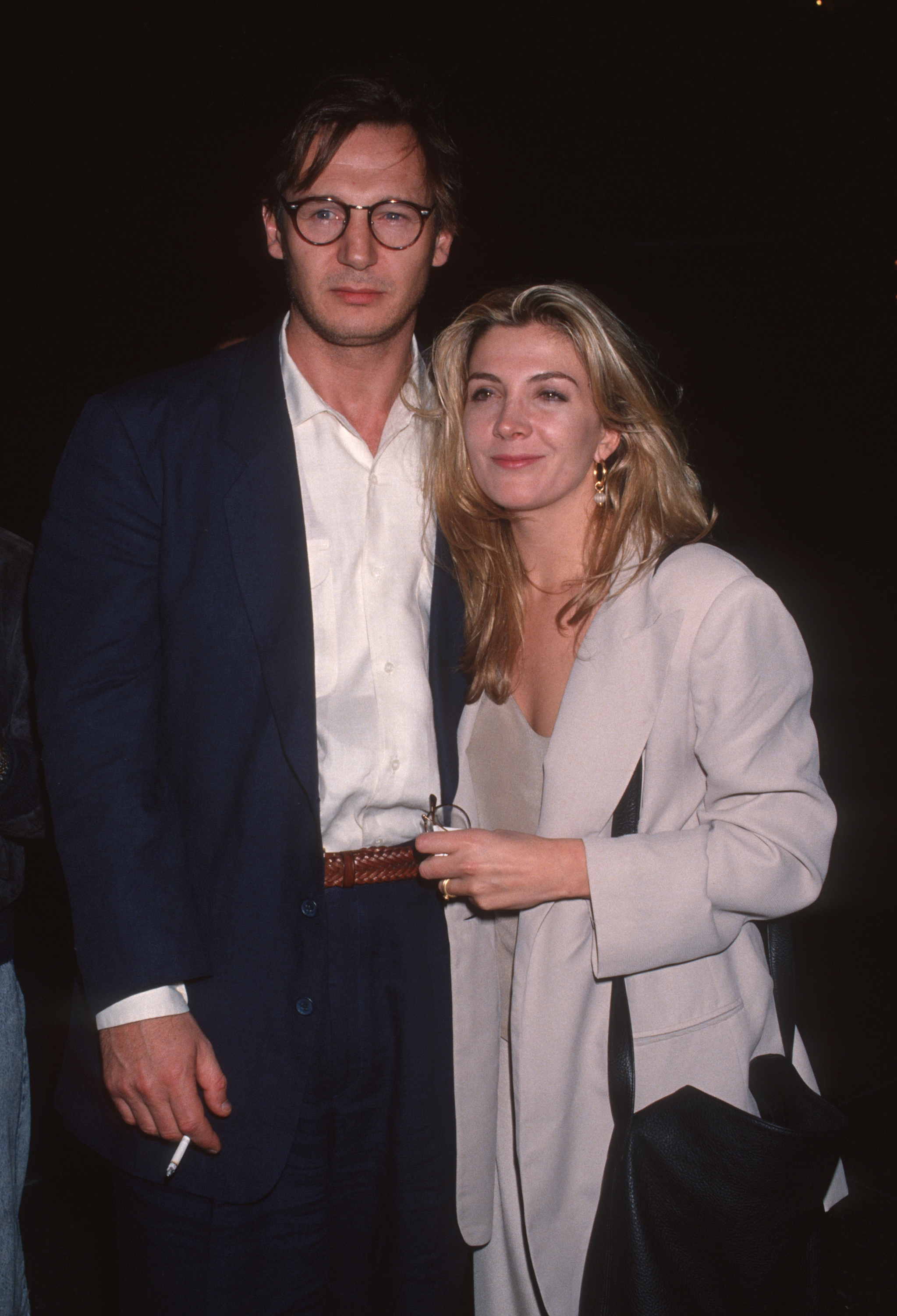Two individuals on a red carpet, one wearing a dark suit and glasses, the other in a light suit and top, smiling and posing together
