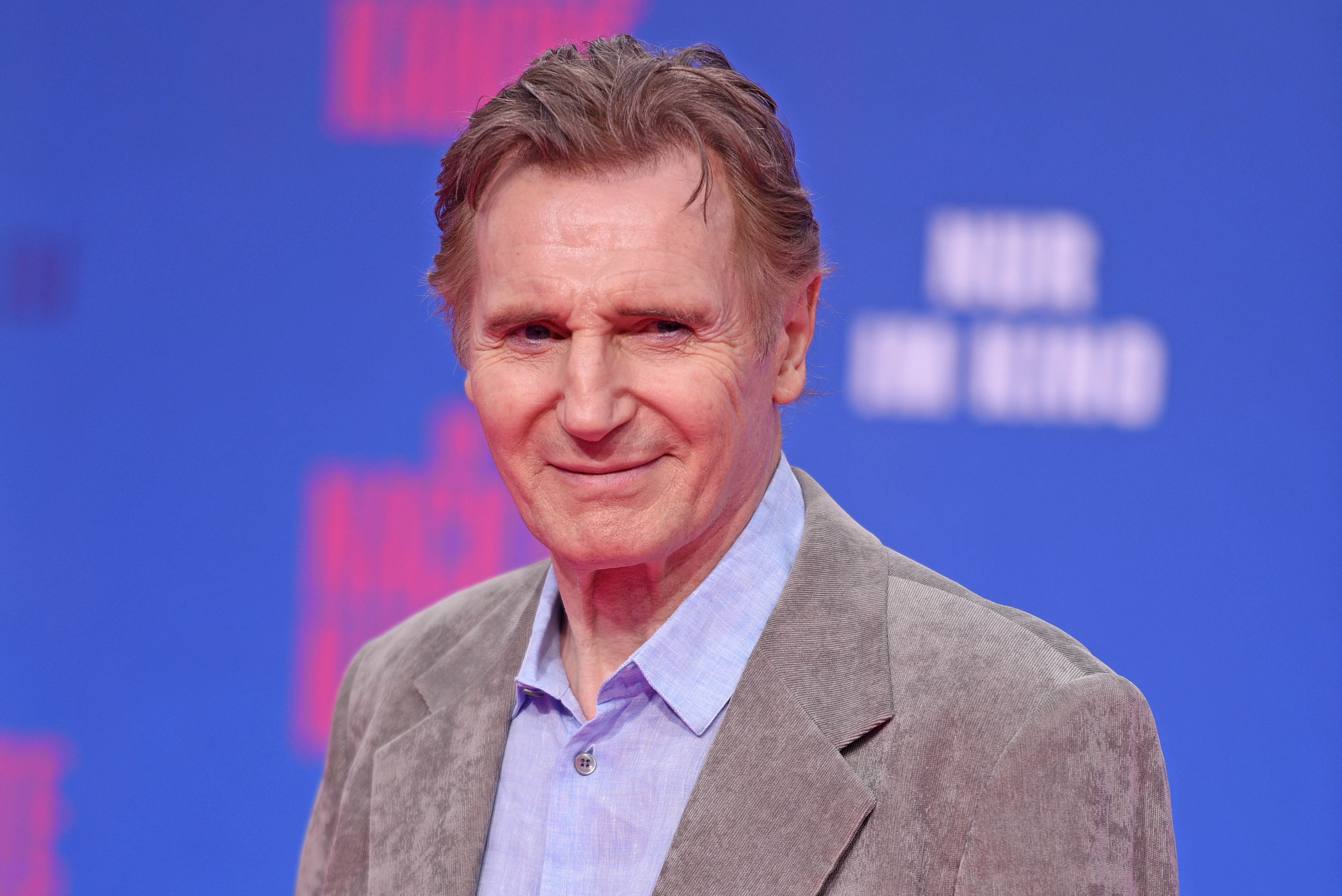 Actor in a light gray blazer and light blue shirt poses on a blue-themed red carpet backdrop with blurred text