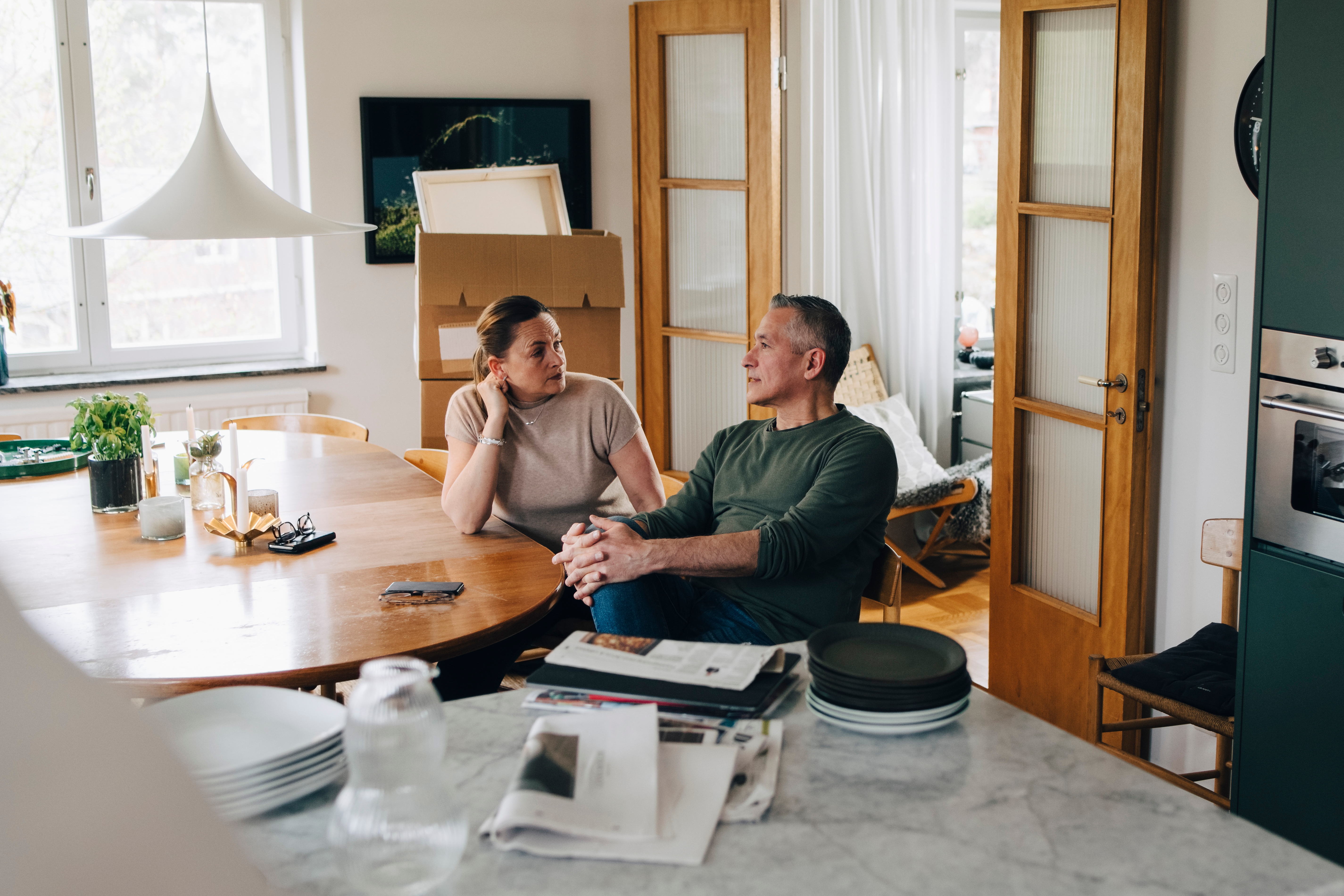 A couple sits talking at a dining table in a cozy, modern kitchen with unpacked boxes around, suggesting moving in or redecorating