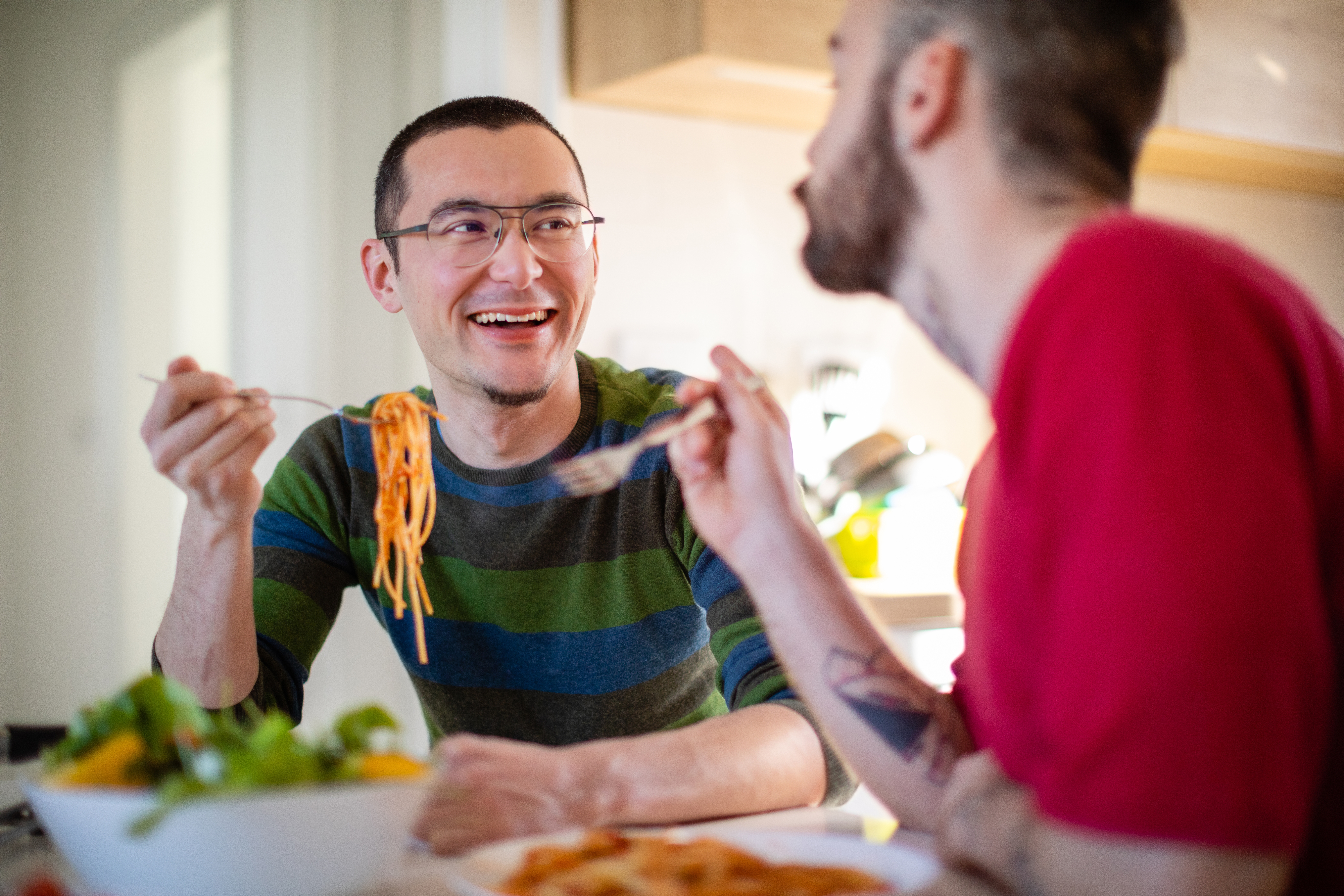 Two people enjoying a meal at home, smiling and sharing pasta and salad, fostering a warm, intimate atmosphere