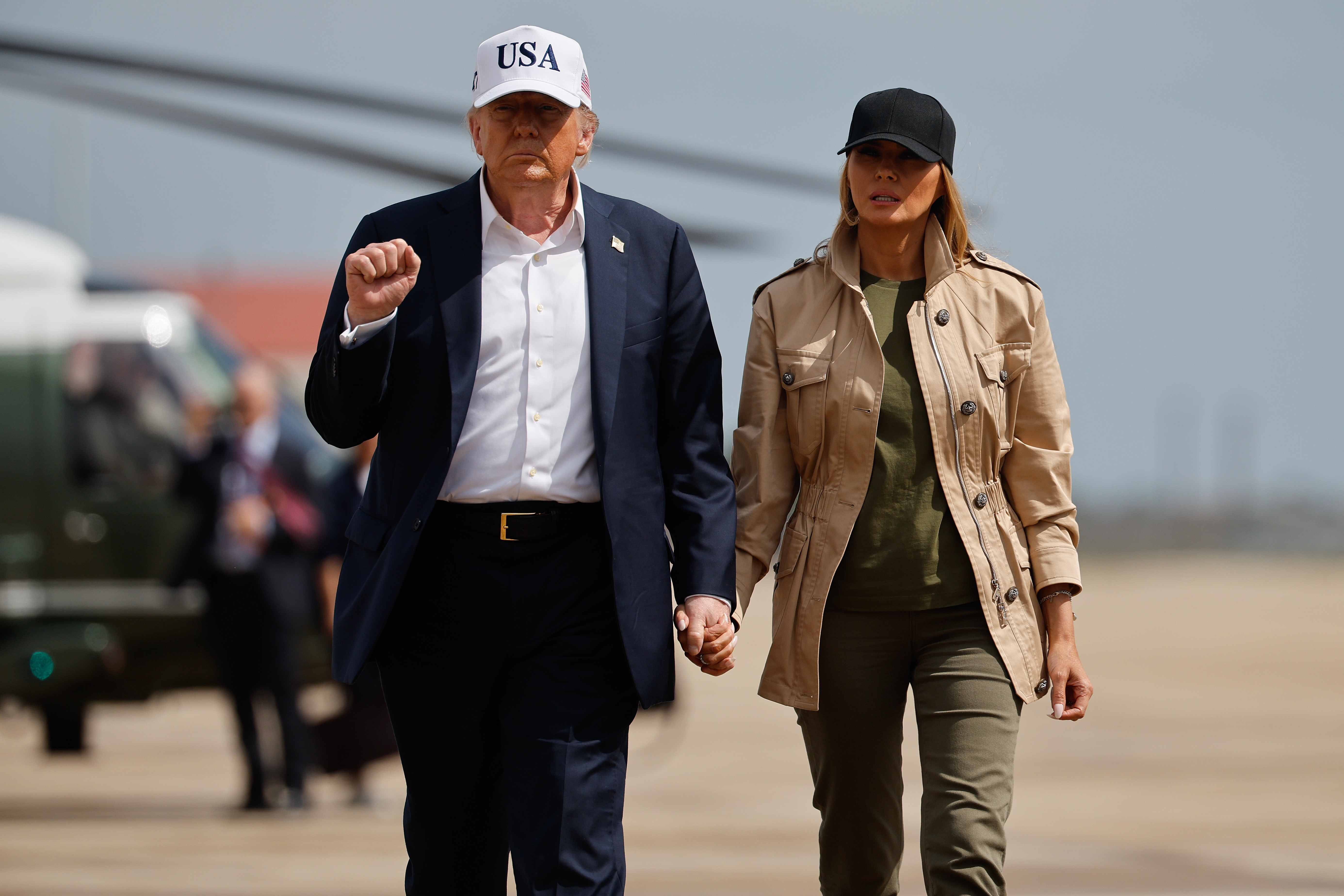 Two individuals walking; one in a suit with a &quot;USA&quot; cap, the other in a casual jacket and cap, holding hands on a tarmac