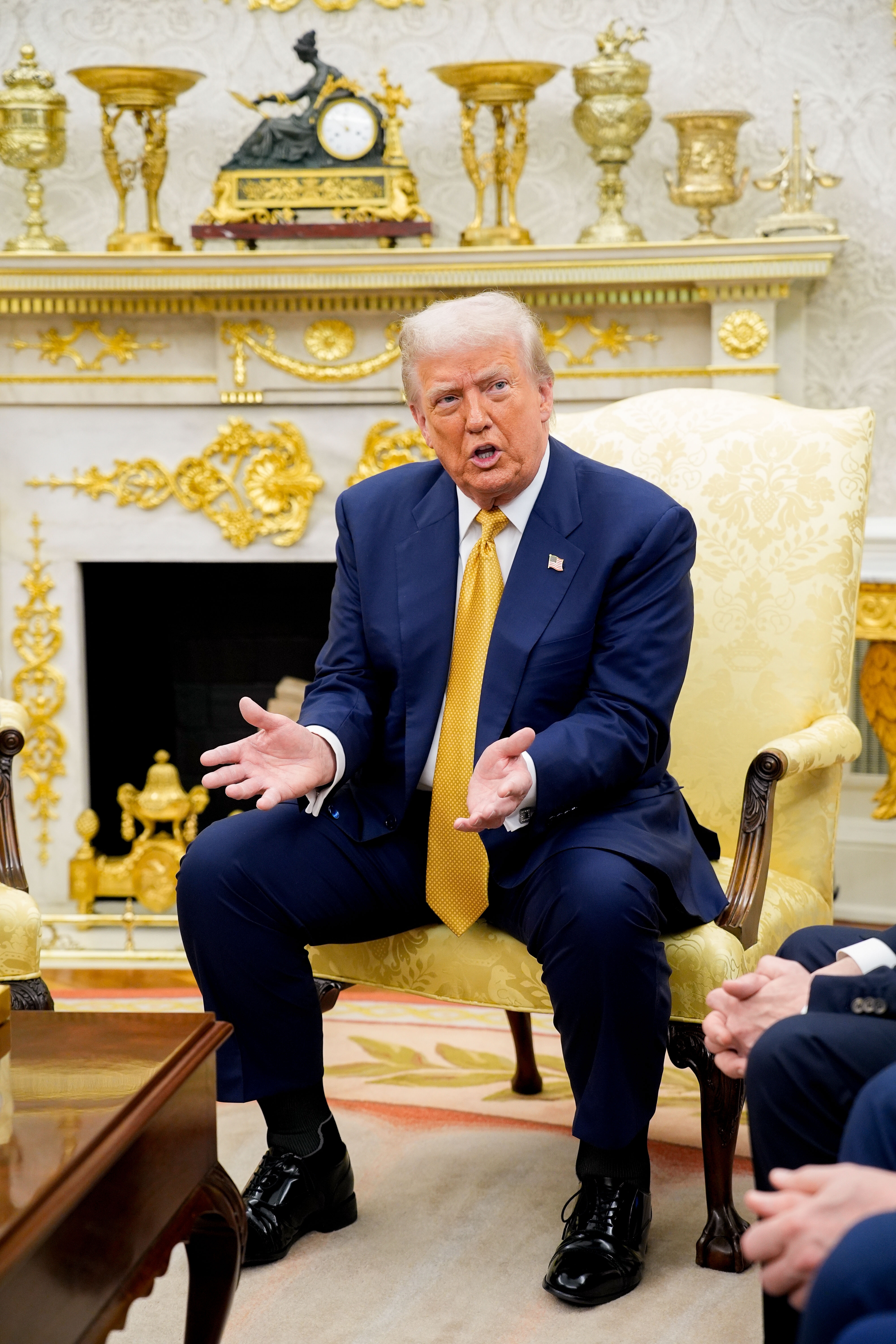 A man in a suit and tie sits in an ornate room, gesturing with his hands, engaged in conversation
