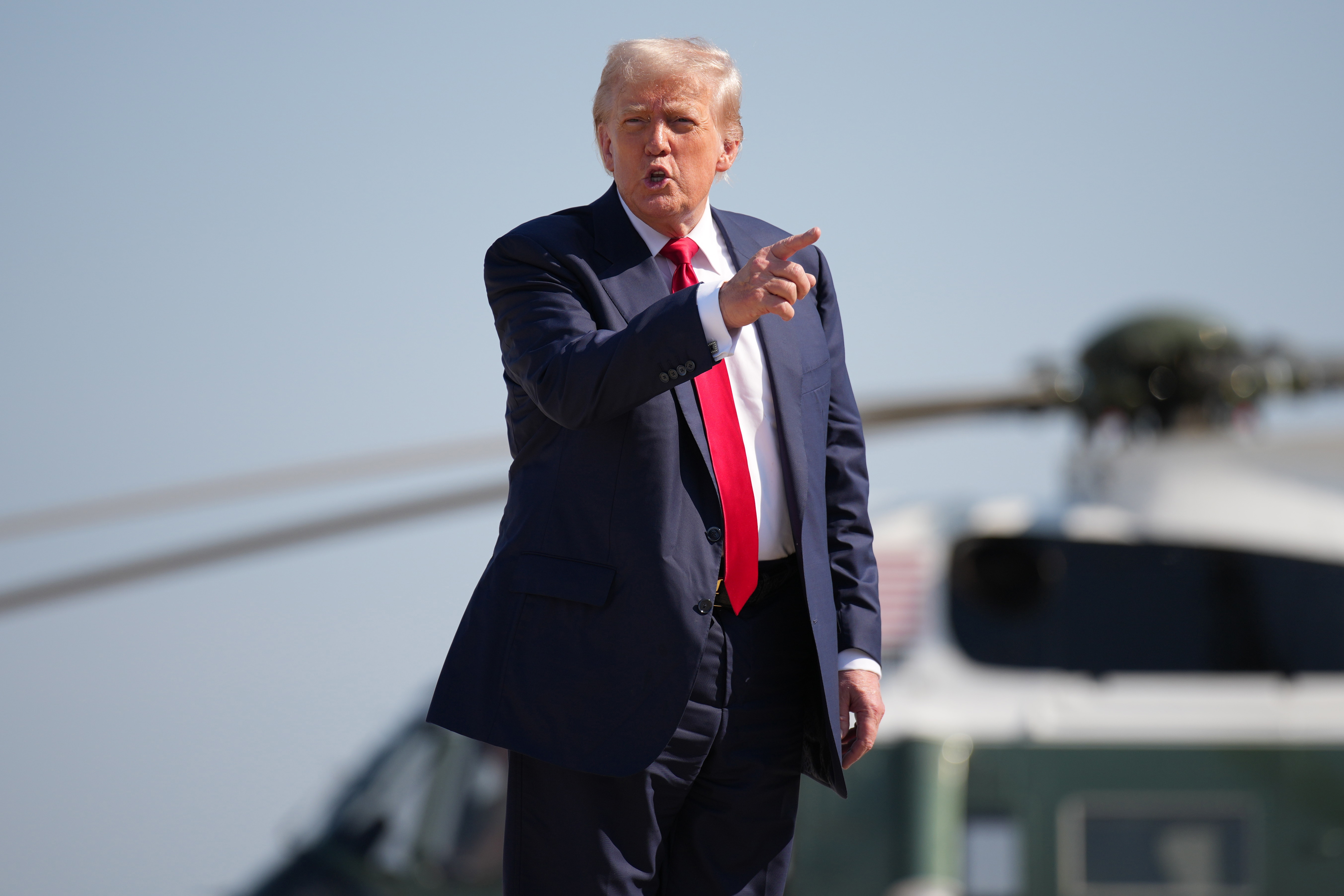 Person in a suit with a red tie gestures outdoors near a helicopter