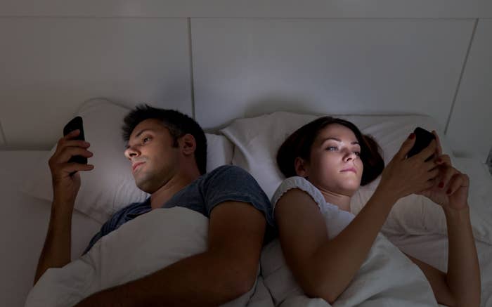 A couple in bed facing away from each other, both focused on their smartphones, suggesting digital distraction in a relationship
