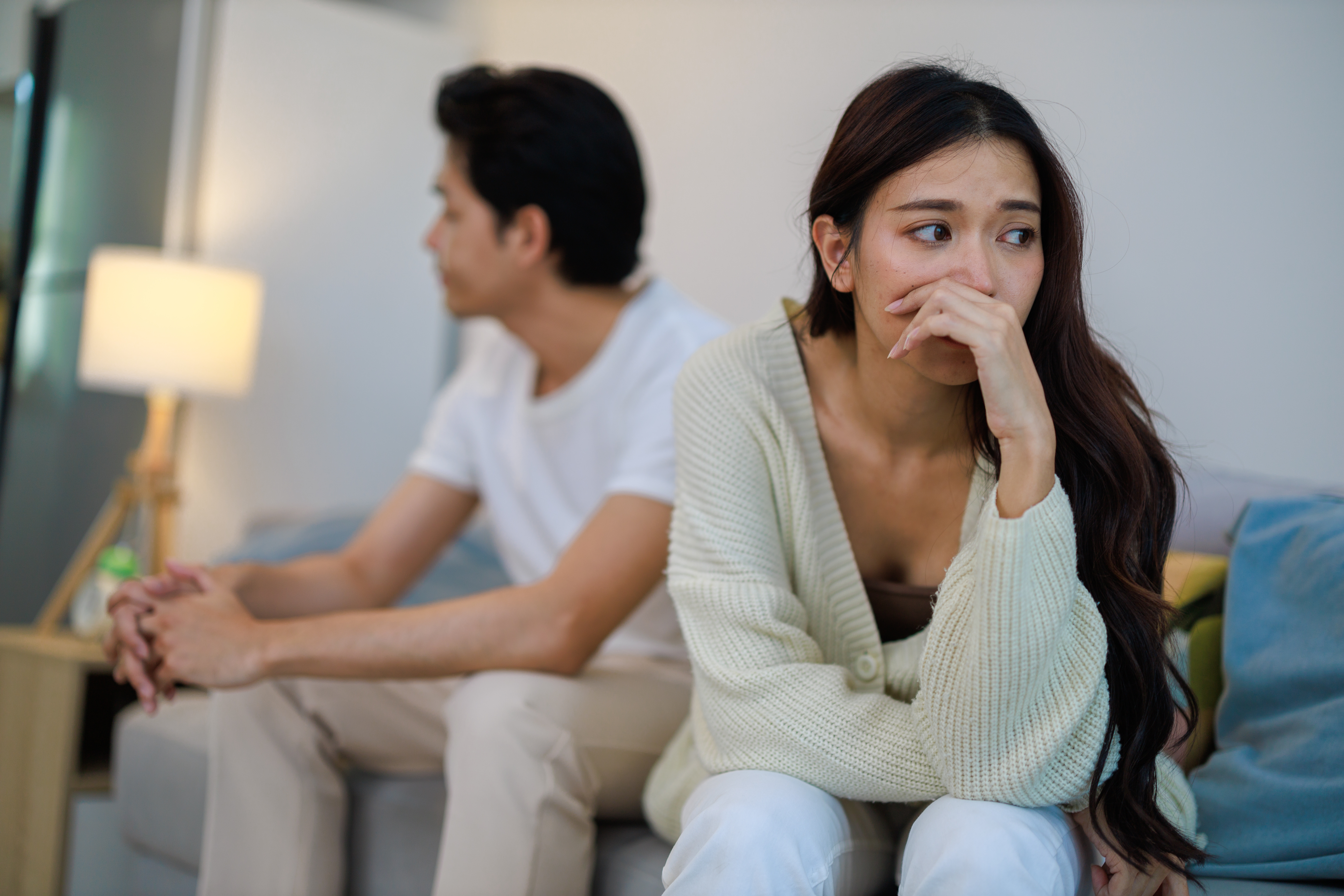 A couple sits on a sofa facing away from each other, looking thoughtful and concerned. The woman has long hair and covers her mouth with her hand