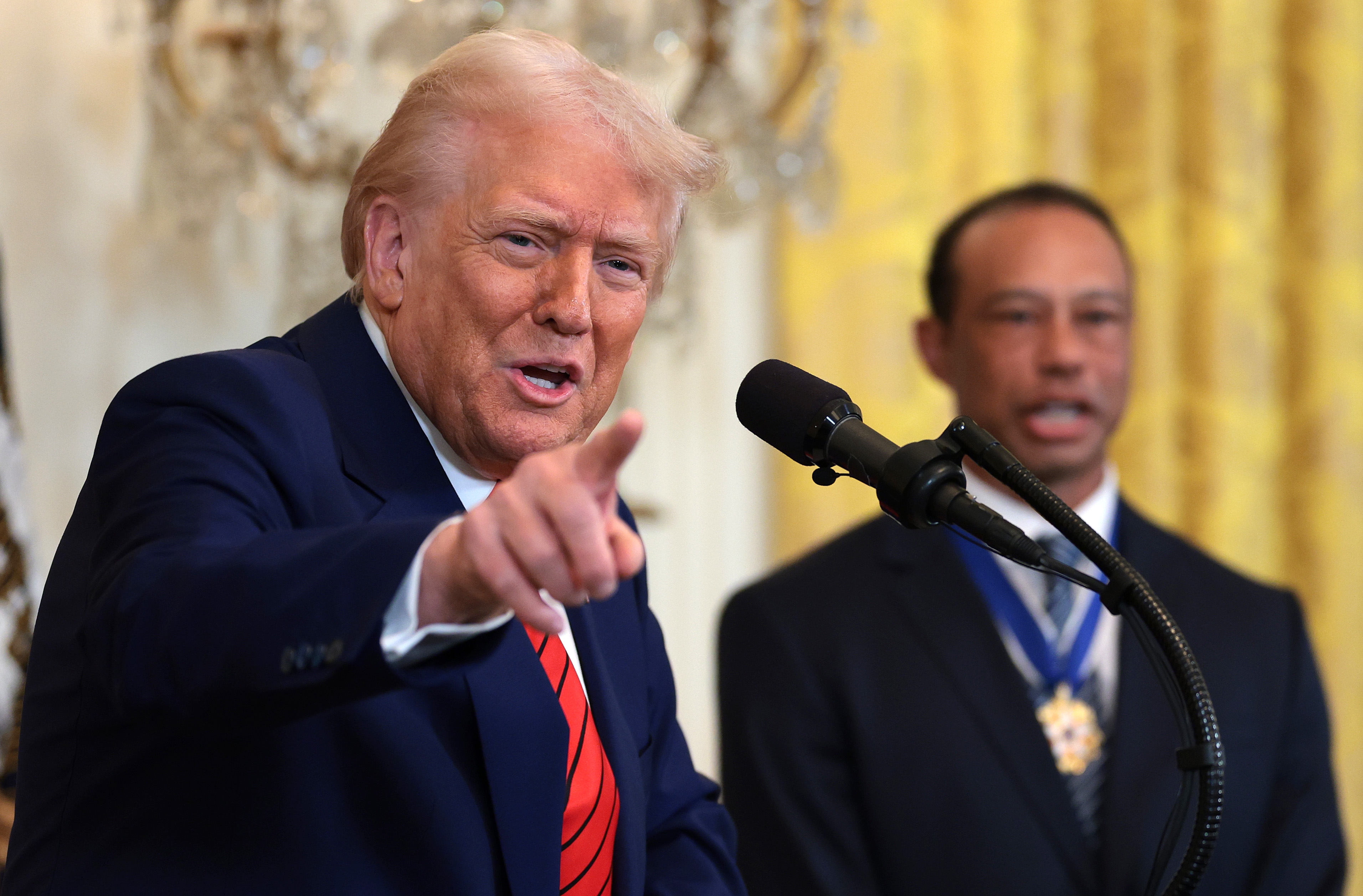 A speaker in a suit points at an audience from a podium, with a person in the background wearing a formal outfit and a visible medal
