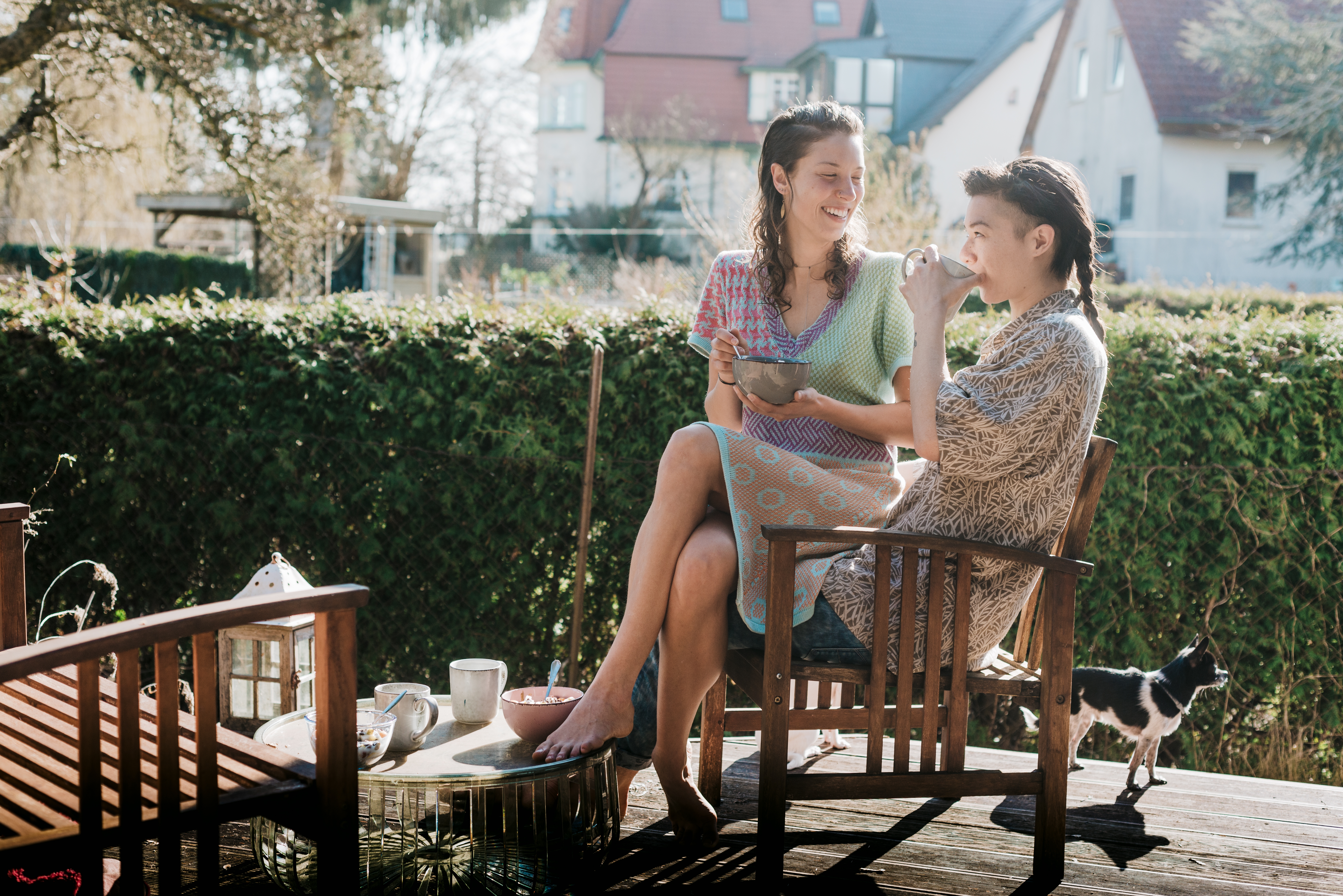Two people enjoy a lighthearted moment on a sunny patio, sharing a snack and smiling at each other. A small dog stands nearby, adding to the serene scene