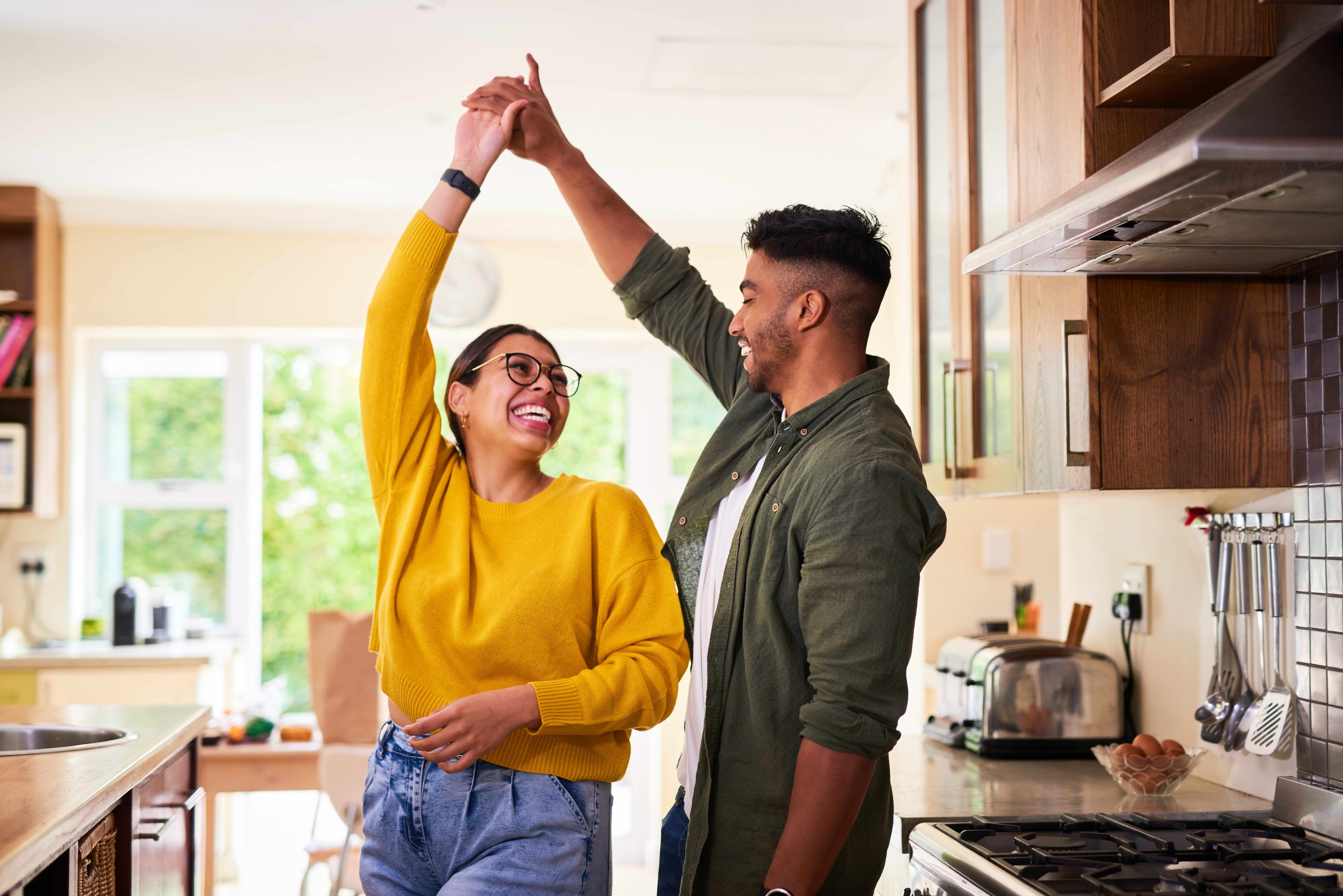 A couple joyfully dances in a kitchen, smiling at each other. The woman wears a sweater and glasses, the man in a casual jacket