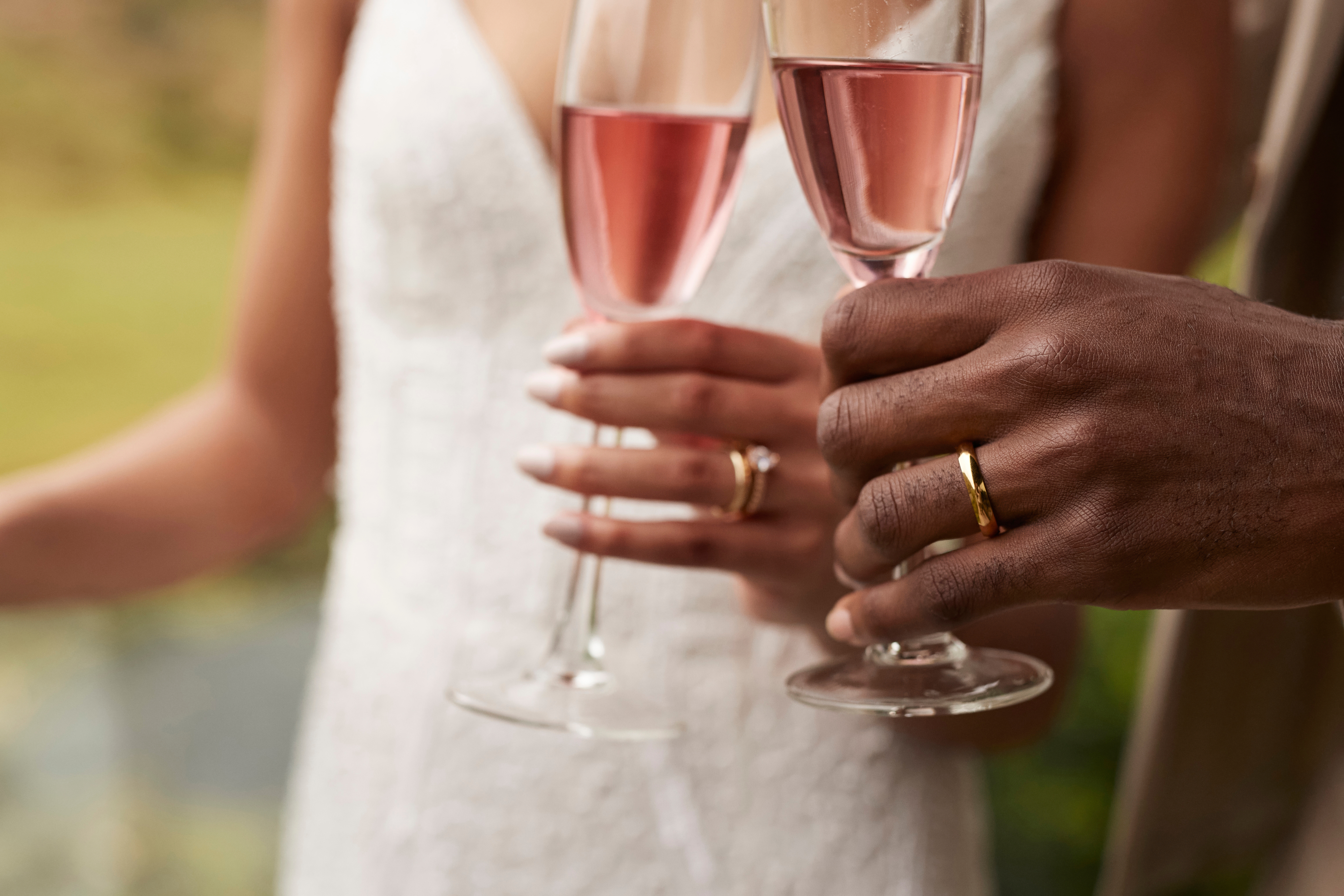 A couple, each wearing a wedding ring, holds champagne flutes filled with pink bubbly, celebrating together
