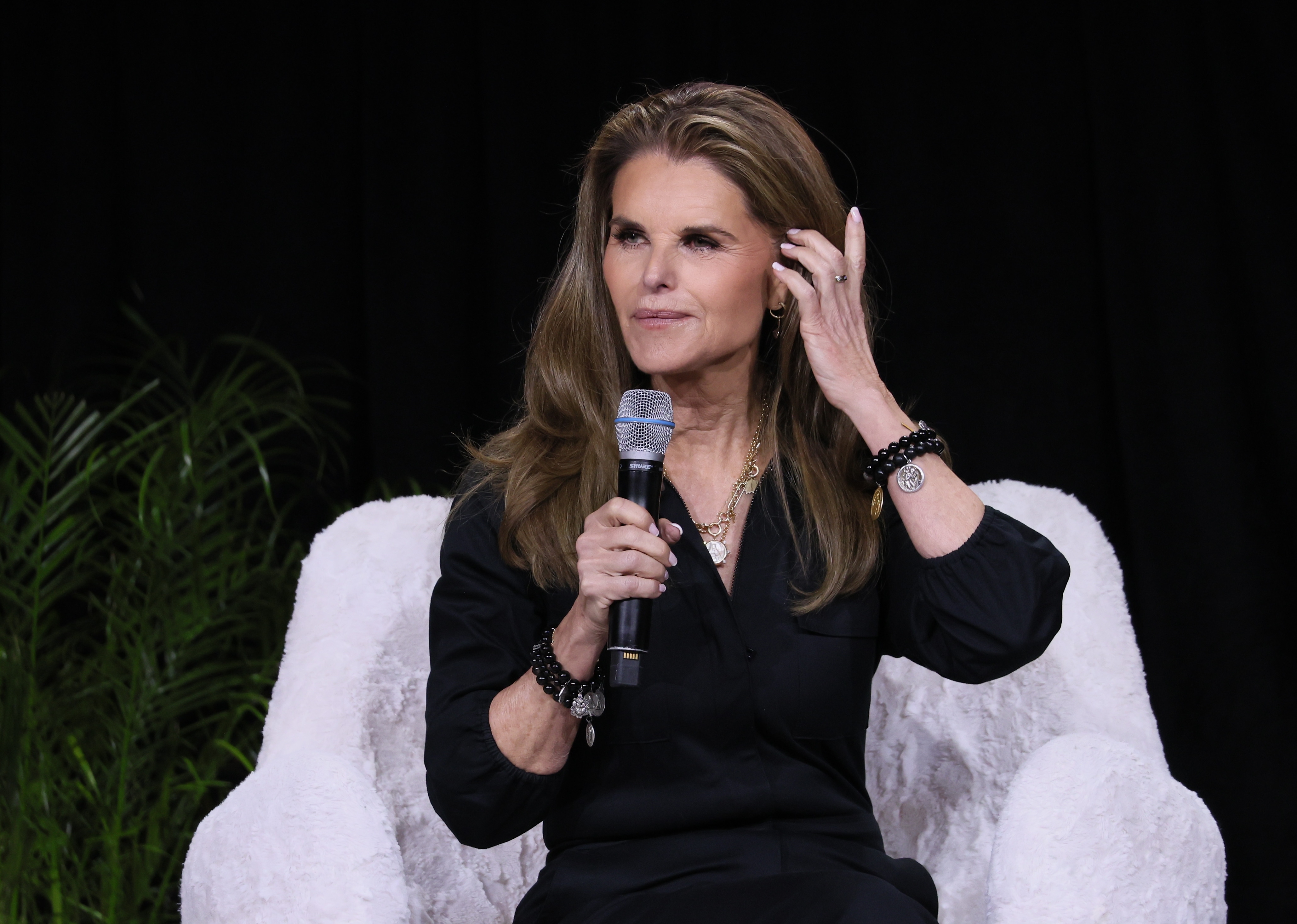 Person seated, holding a microphone, wearing a stylish black outfit, gesturing thoughtfully during an event