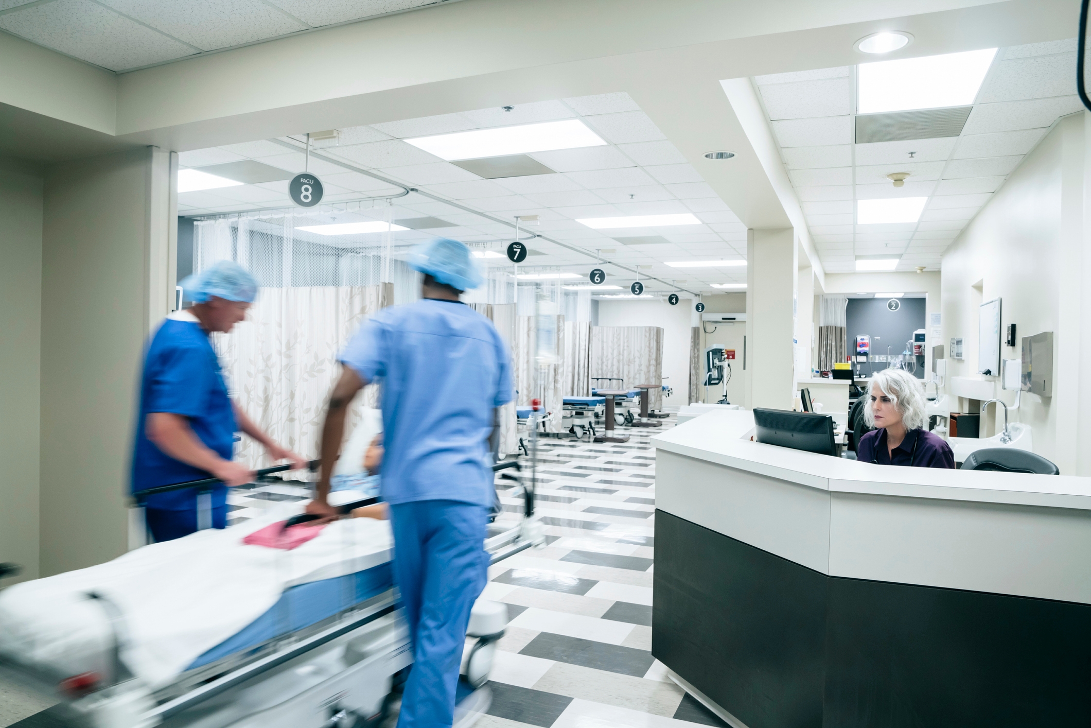 Hospital staff move a patient on a gurney past a reception desk in a busy hospital corridor
