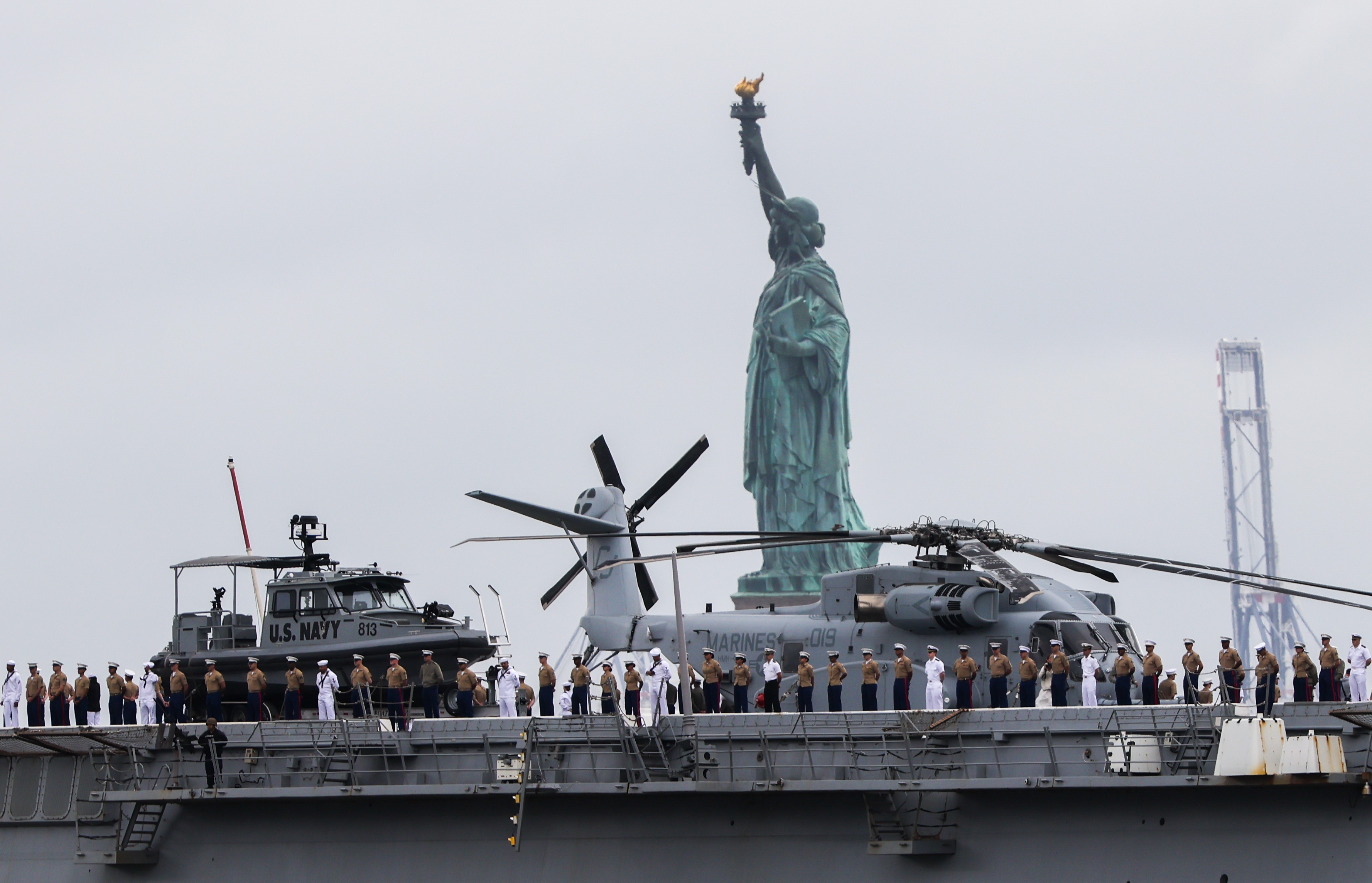 Sailors in uniform stand on the deck of a Navy ship with helicopters and a boat, passing by the Statue of Liberty