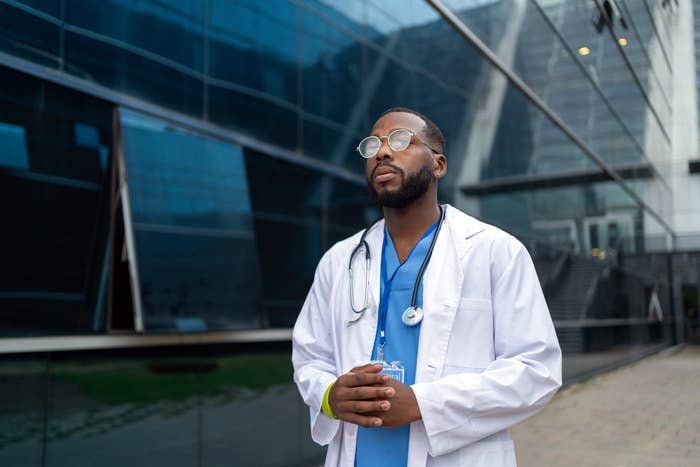 A doctor in blue scrubs and a white coat stands confidently in front of a modern glass building, wearing glasses and a stethoscope