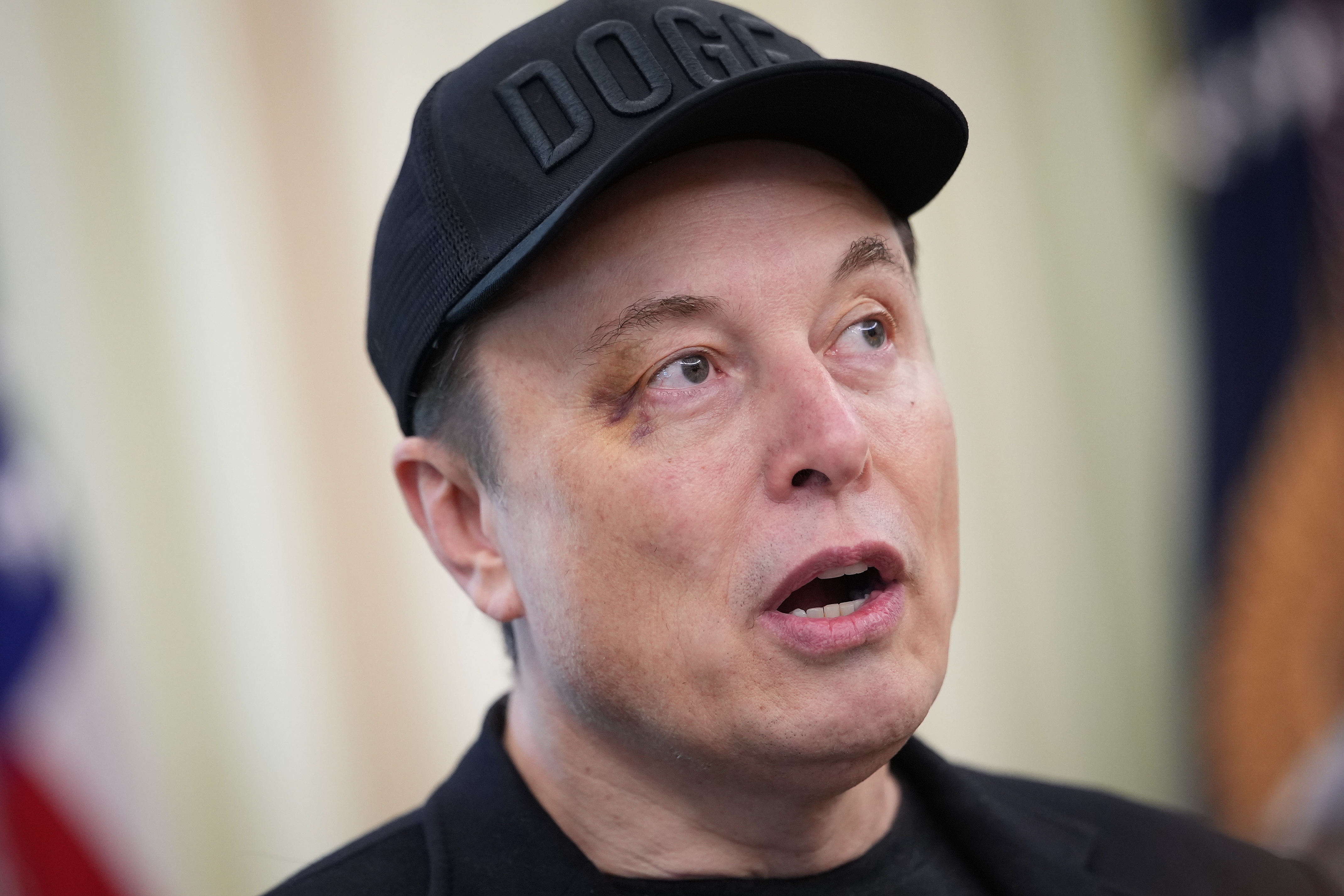 Person wearing a &quot;DOGE&quot; cap speaking, mid-sentence, with a slight bruise visible under one eye