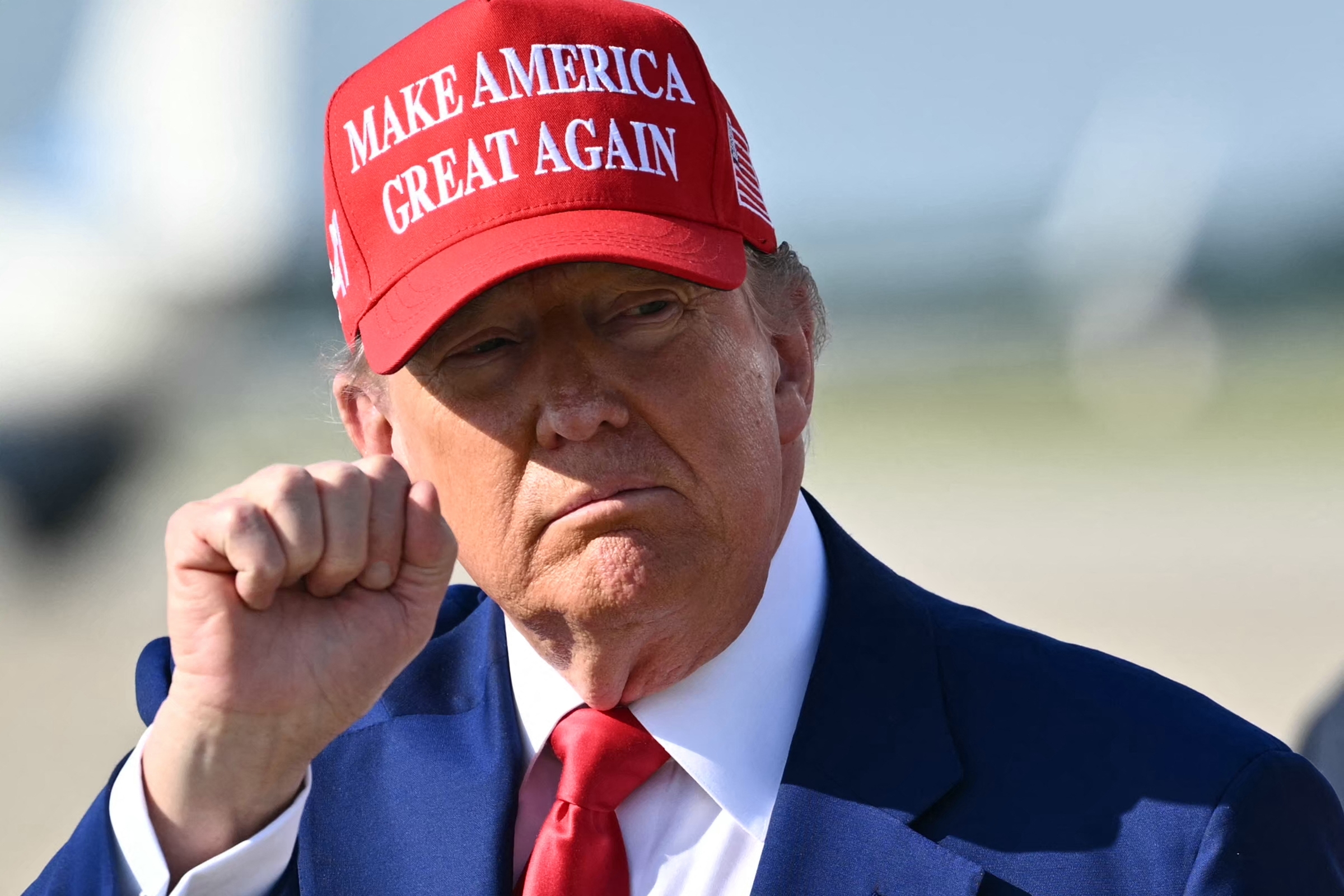 Man wearing a &quot;Make America Great Again&quot; cap, blue suit, and red tie, raises his fist outdoors