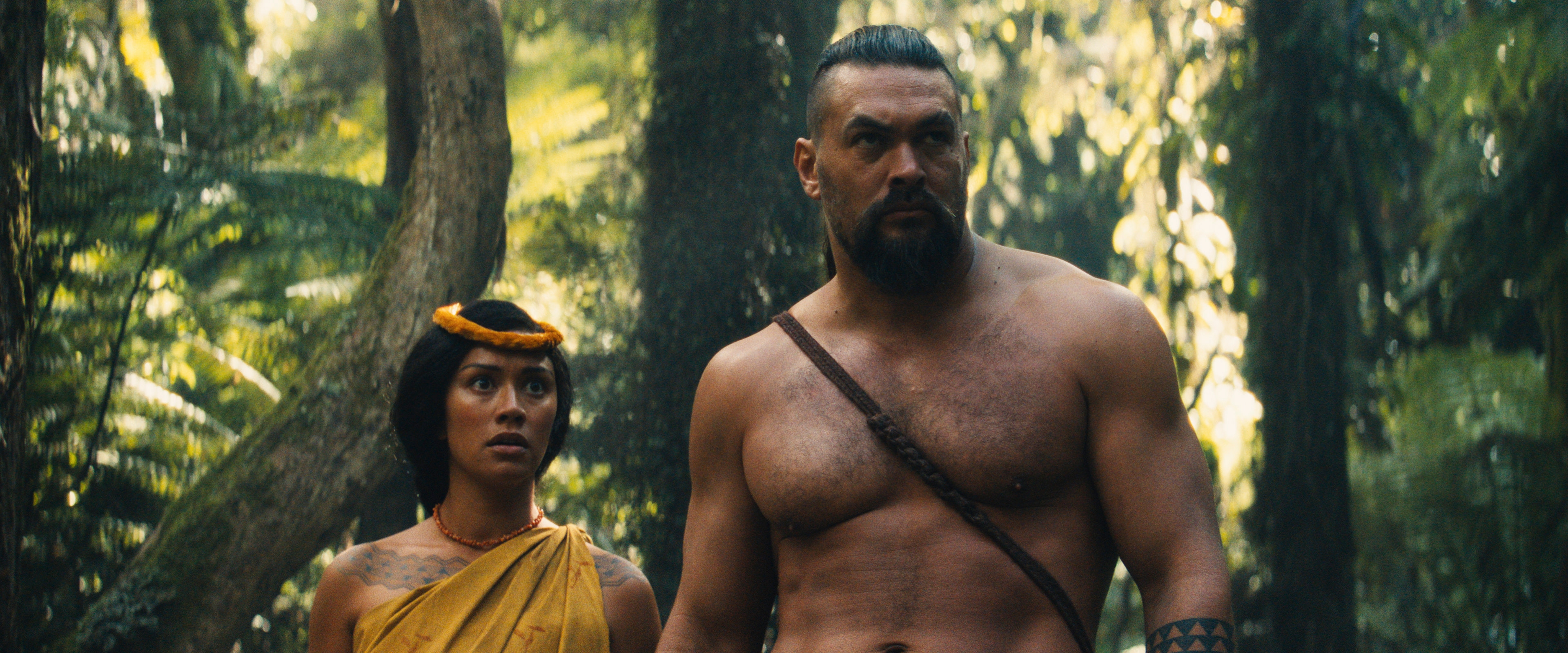Two people in a jungle setting, one wearing a strapless garment and headband, the other shirtless with a crossbody strap, look intently beyond
