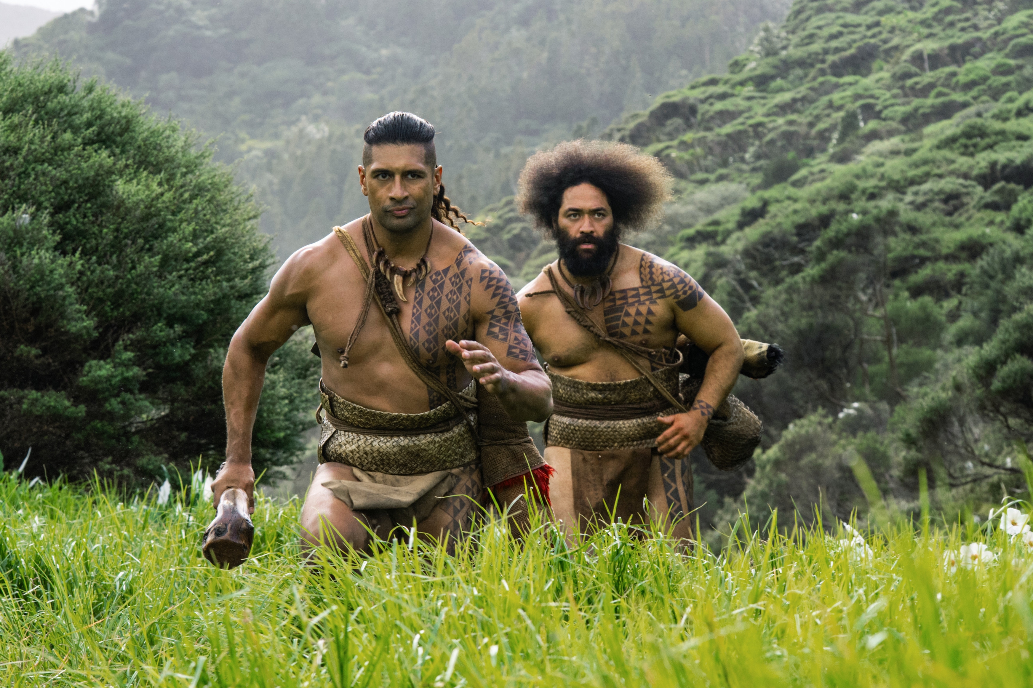 Two men with traditional tattoos run through a lush landscape, wearing woven waist garments. They carry traditional items, evoking historical cultural imagery