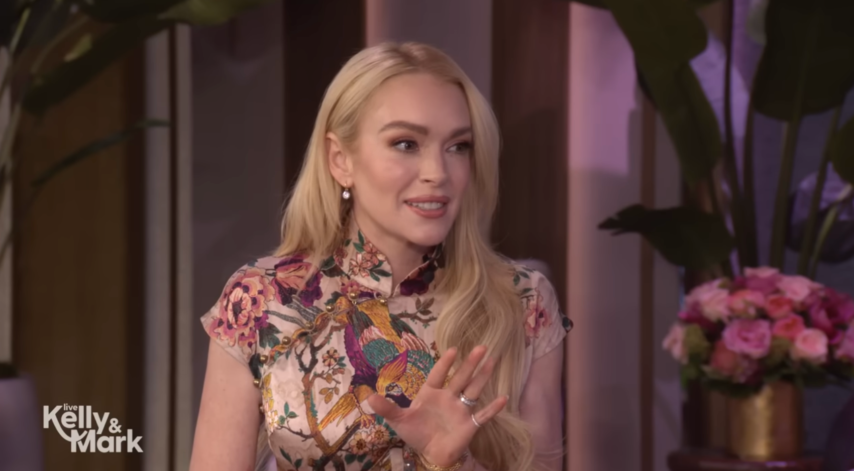 Lindsay Lohan with long blonde hair wearing a floral-patterned outfit is gesturing while sitting in a talk show studio
