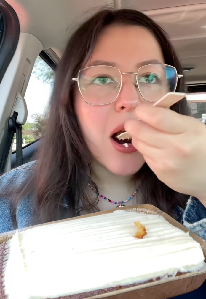 Person in car eating a slice of cake with a fork. Wearing glasses and a beaded necklace