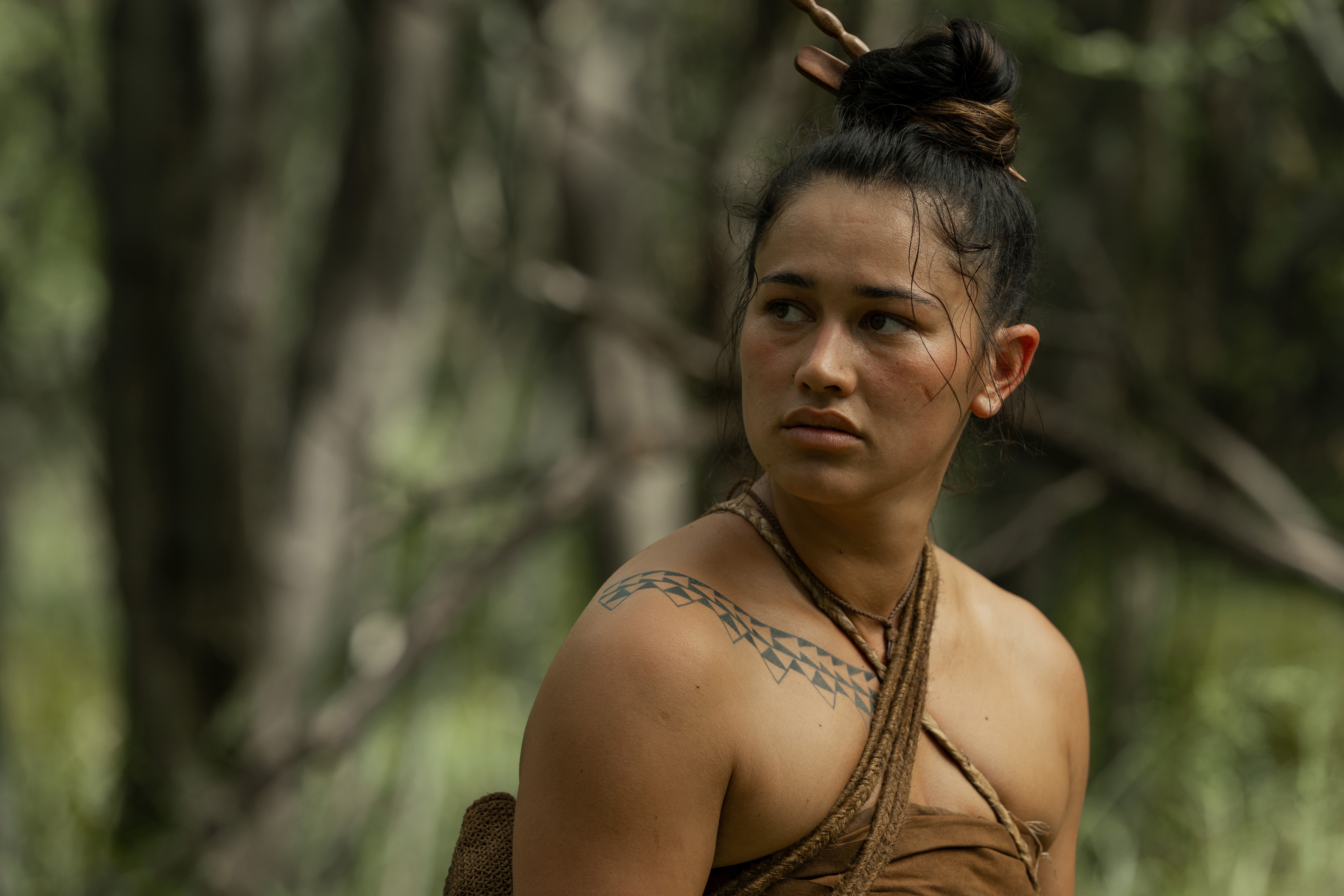 Person outdoors wearing tribal-inspired attire with a tattoo on their shoulder, looking to the side with trees in the background
