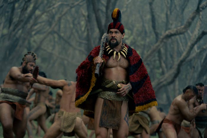 A person dressed in traditional warrior attire with a headpiece and fur robe walks through a group of similarly dressed individuals in a forest setting