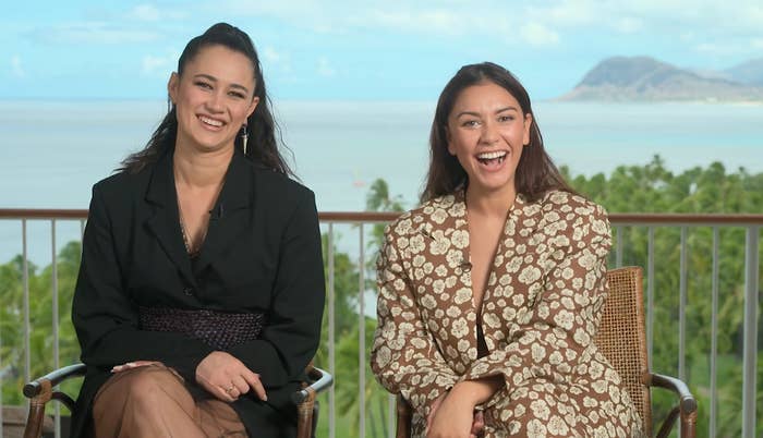 Two people sit and smile outdoors, with a scenic ocean background. One wears a black outfit, and the other wears a patterned dress