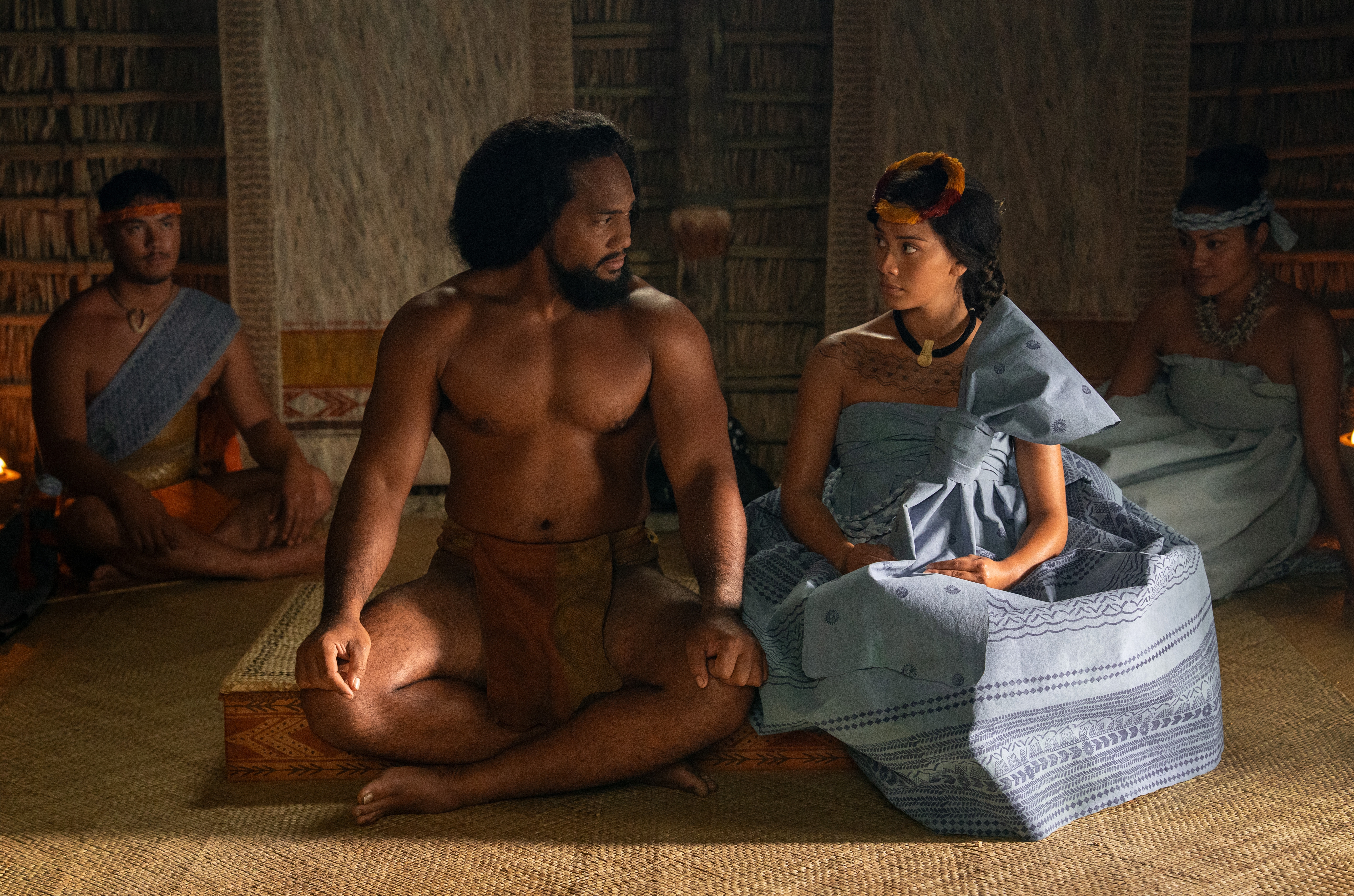 A group of people in traditional Pacific Island attire sit inside a wooden structure, engaged in a serious conversation