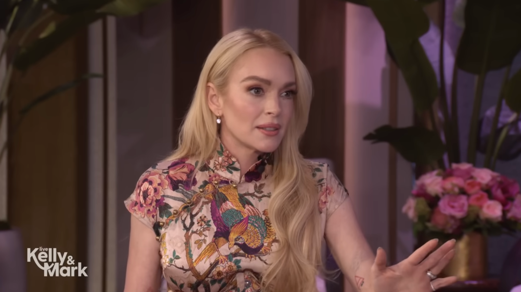 Lindsay Lohan with long blonde hair is sitting in an interview setting, wearing a floral patterned top, gesturing with one hand