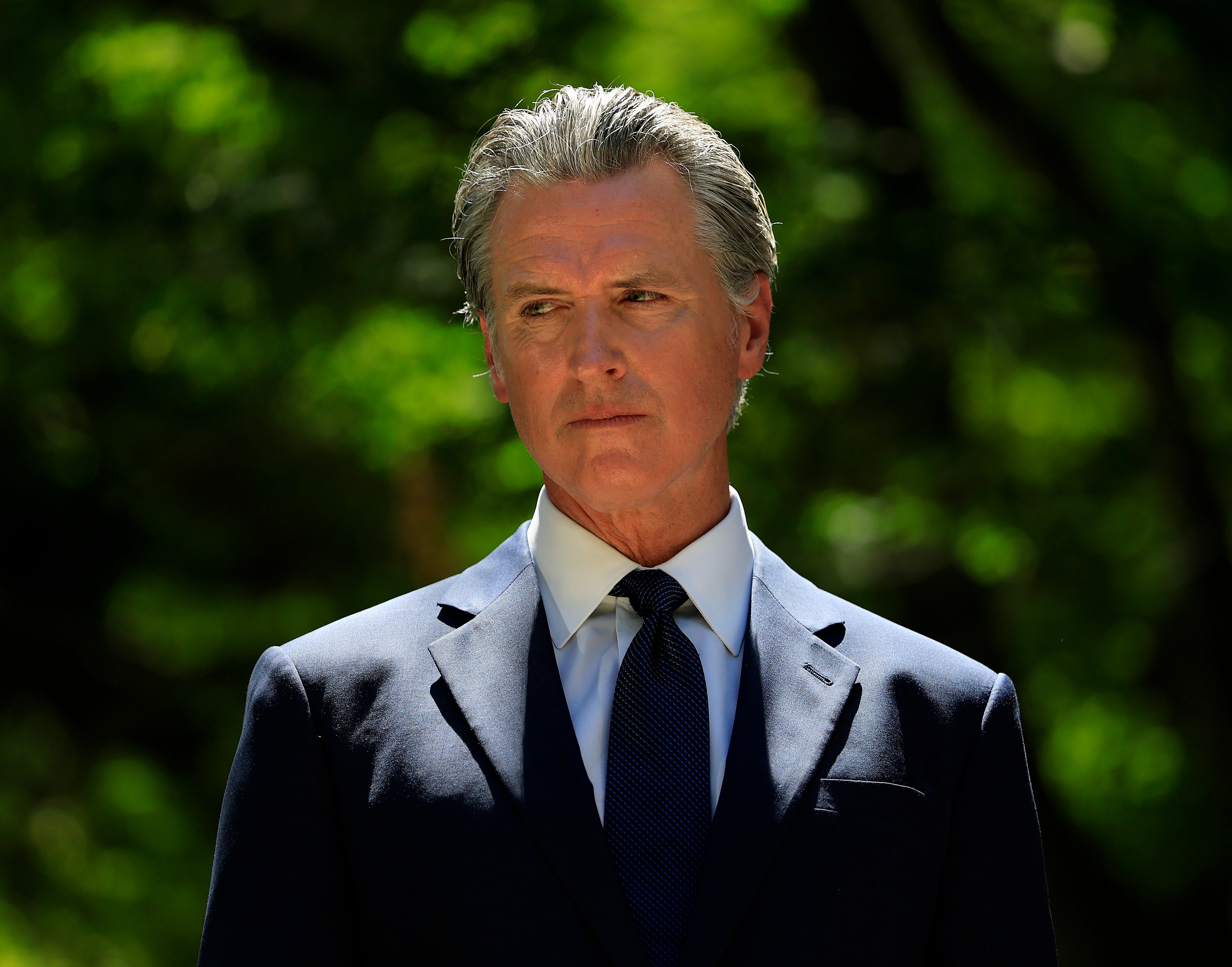 A man in a dark suit and tie stands outside, looking serious, with a blurred natural background