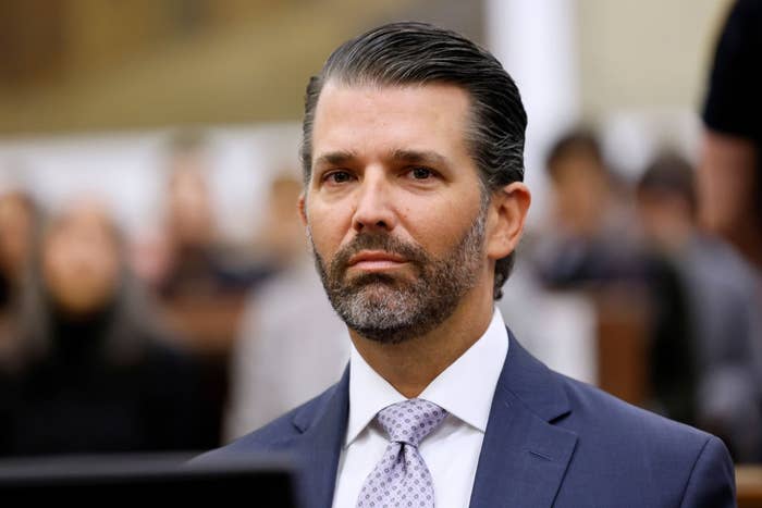 A person in a suit and tie is seated in what appears to be a formal setting, possibly a courtroom or legislative hearing