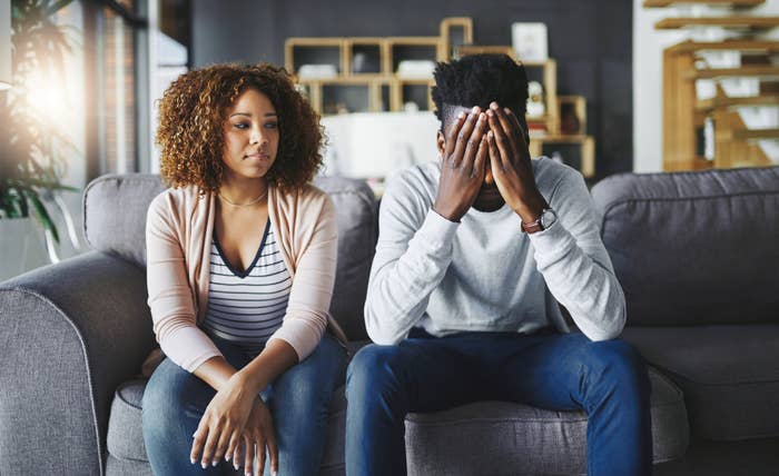 A woman sits on a couch looking concerned while a man beside her covers his face with his hands in apparent distress