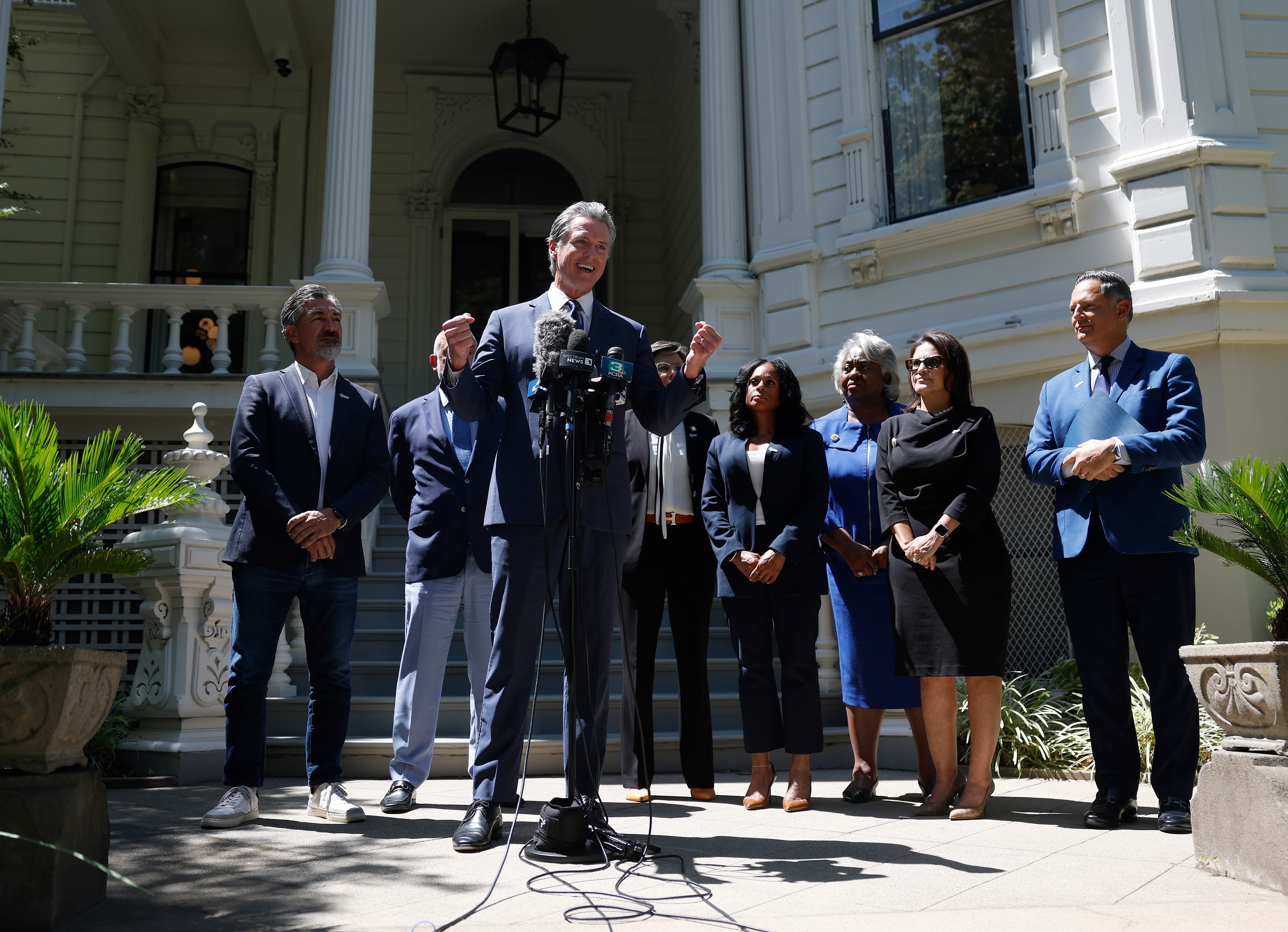 Group of officials standing on a building&#x27;s steps, with a central figure in a suit speaking at a podium with microphones